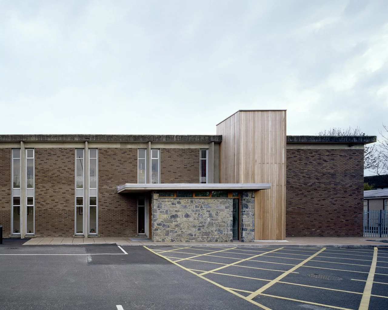 Modern brick building with tall, narrow windows, a stone wall feature, and a wooden vertical structure against the wall. An empty parking lot with yellow-striped spaces in front.