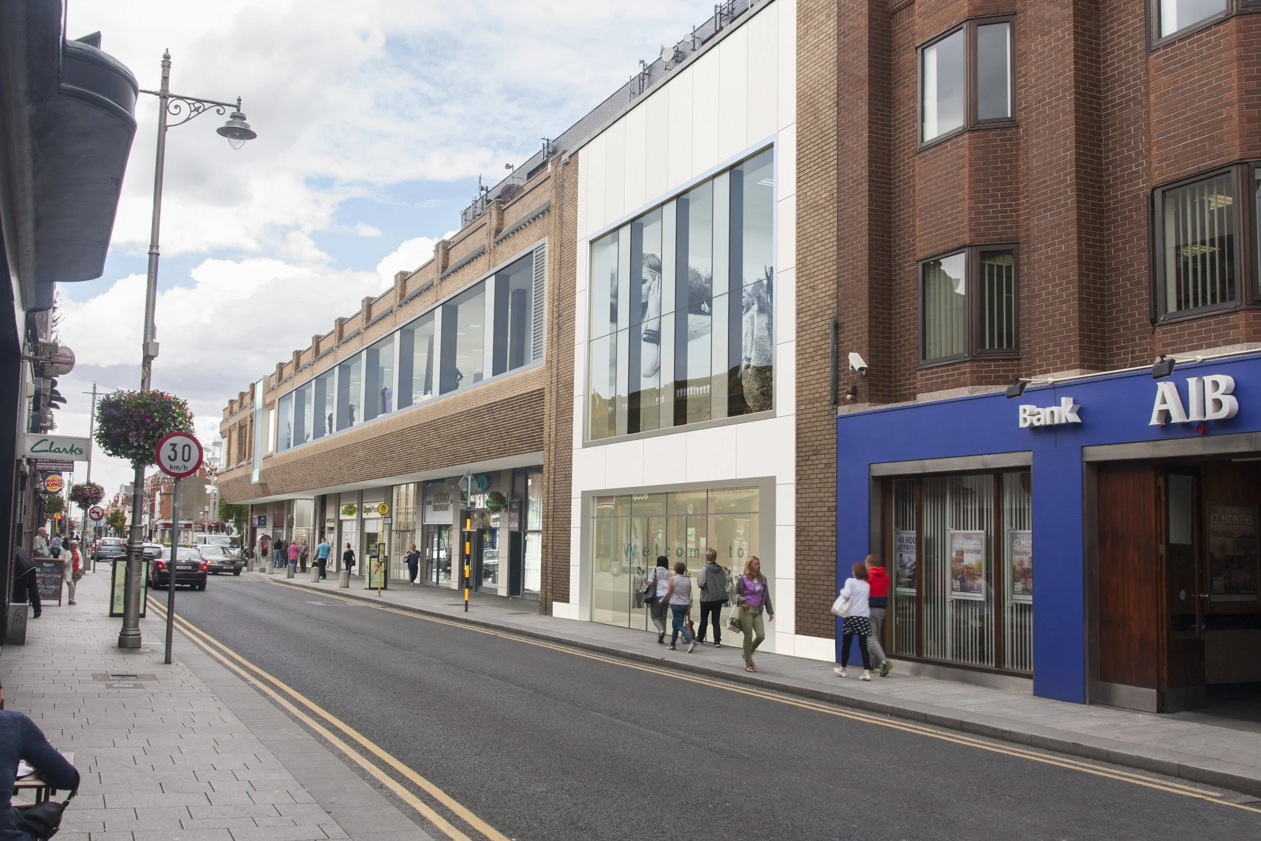 City street with shops, pedestrians walking, and cars parked along the sidewalk. Modern buildings with large windows, including a bank with blue signage, and hanging flower baskets. Overcast sky.