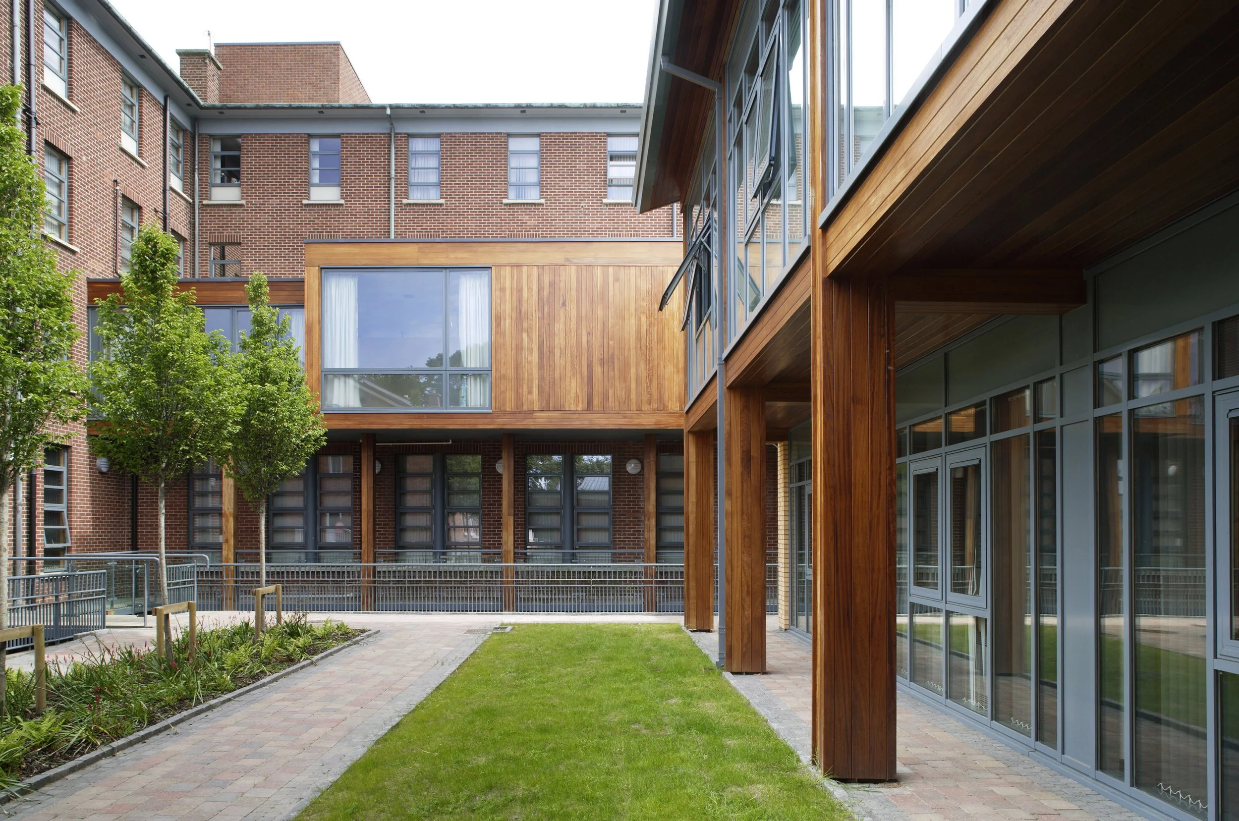 Modern apartment building with brick and wooden exterior, large windows, and a small landscaped courtyard with a paved walkway, grass, and trees.