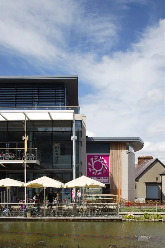 Modern building with glass exterior, outdoor seating area with umbrella, and a pink and white sign for the Mill Theatre beside a river.