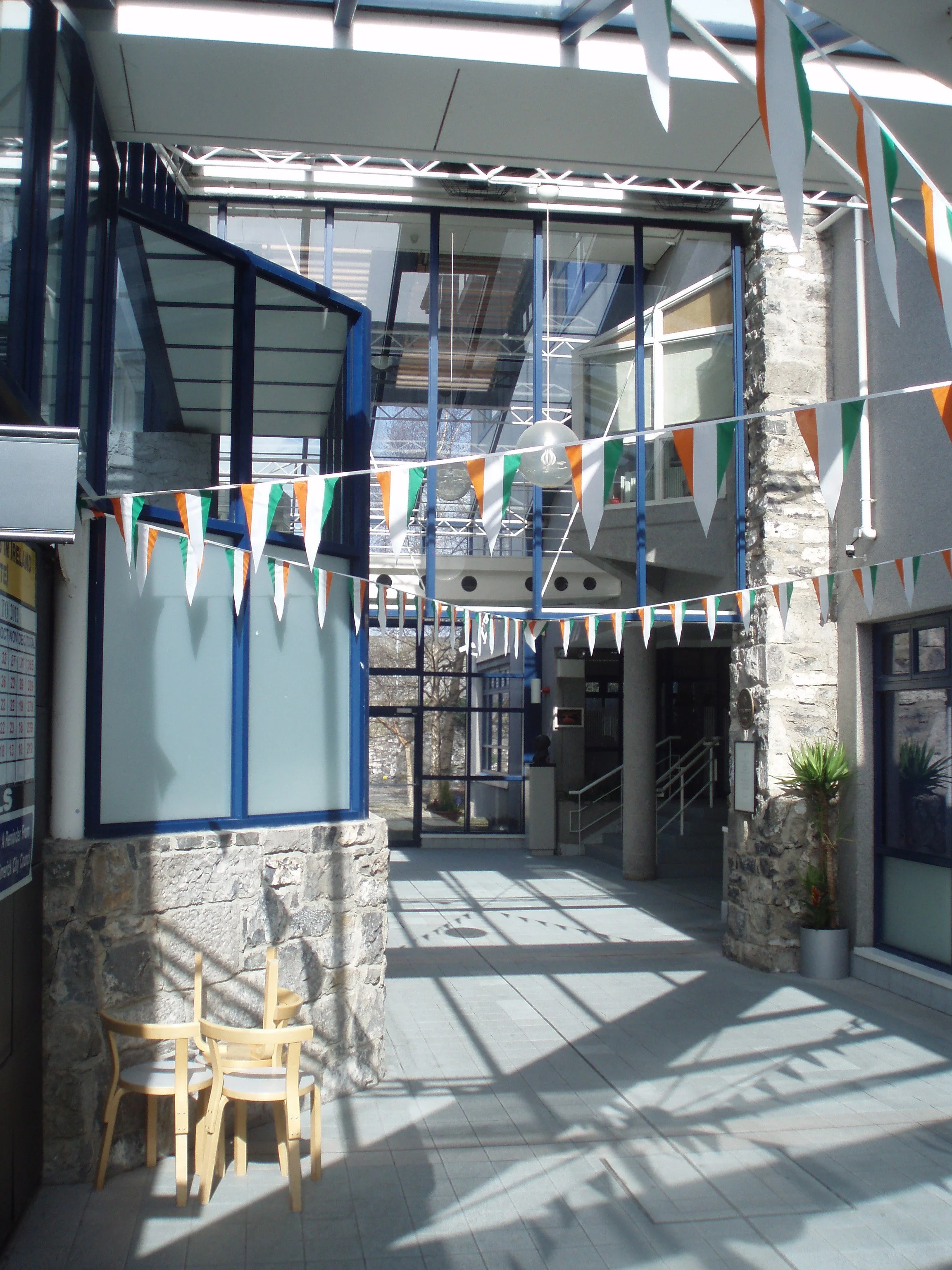 Outdoor courtyard decorated with triangular flags in orange, white, and green, with modern glass and stone building in the background.