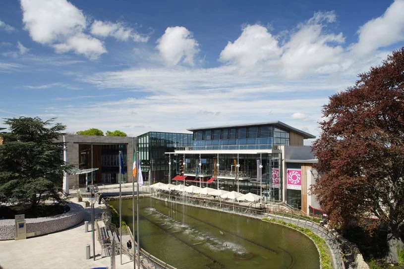 Modern glass building with outdoor patio and water fountain, trees, and cloudy sky.