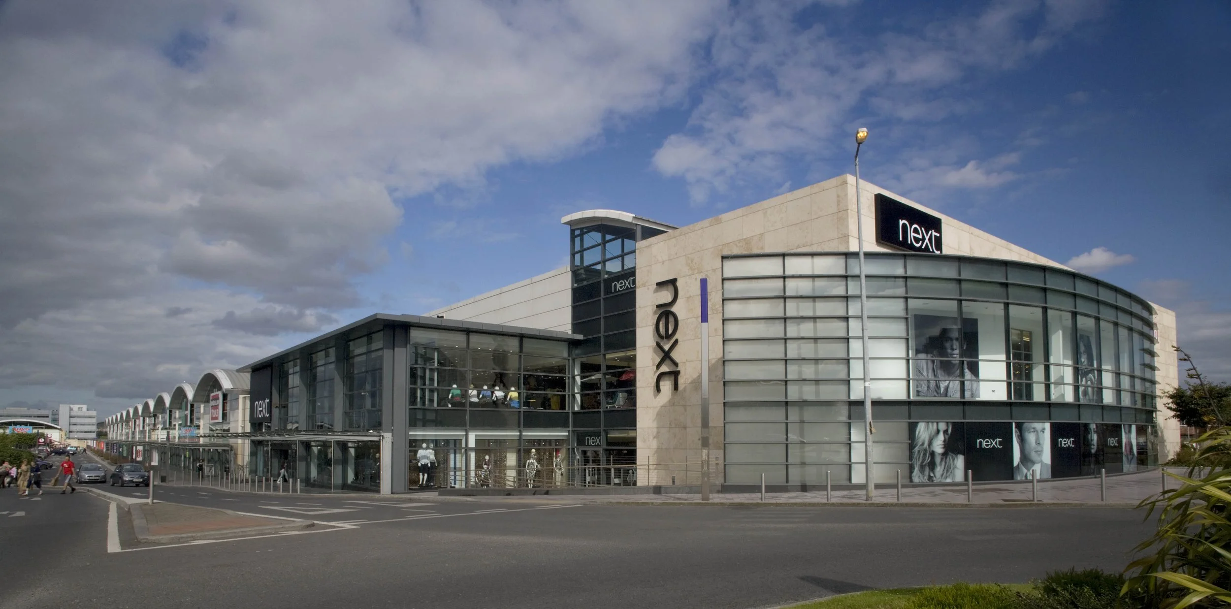 Exterior view of a modern shopping mall with a large glass facade, labeled 'next', under a partly cloudy sky.