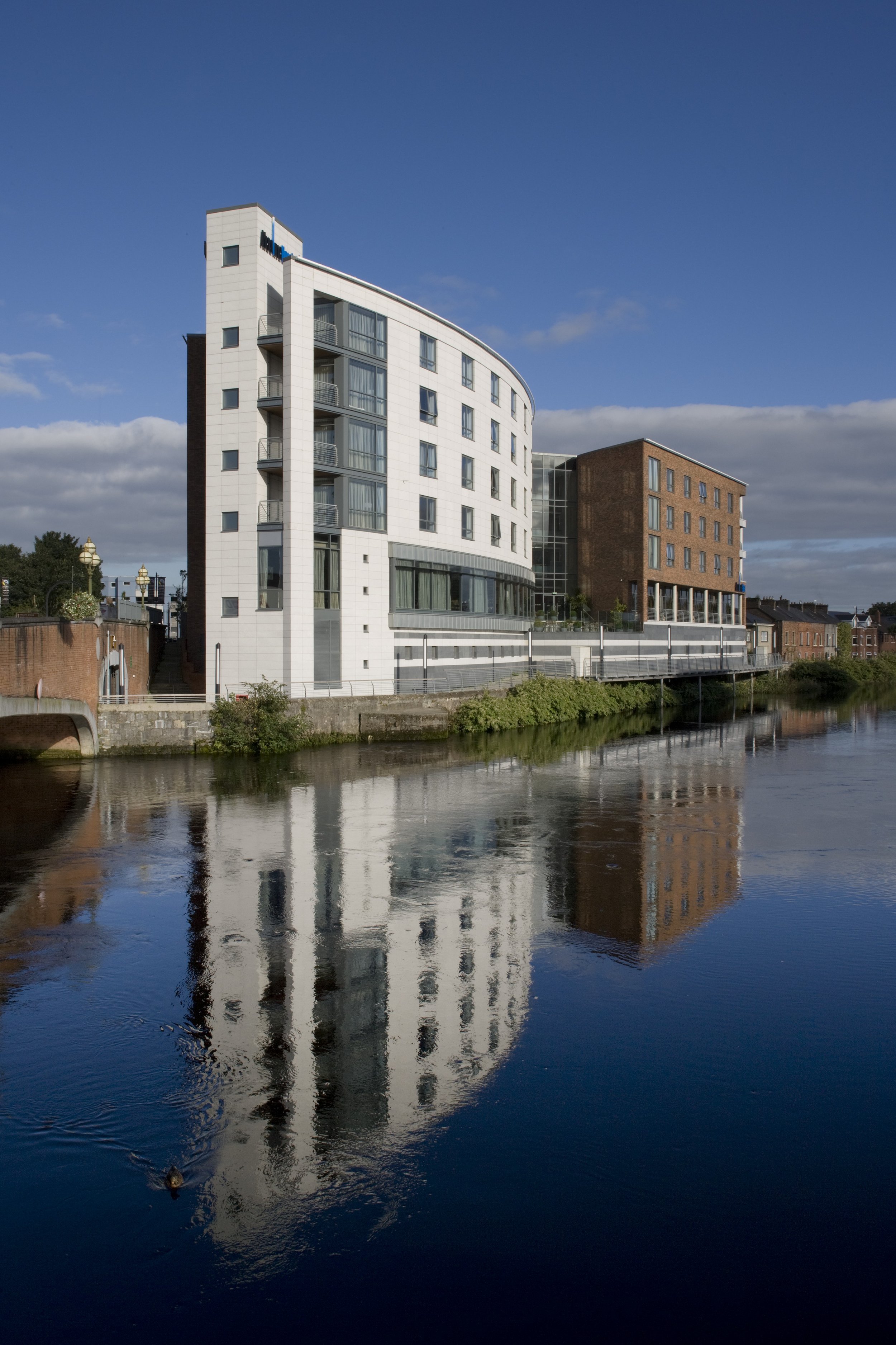 Modern multi-story buildings reflecting on a river under a partly cloudy sky.