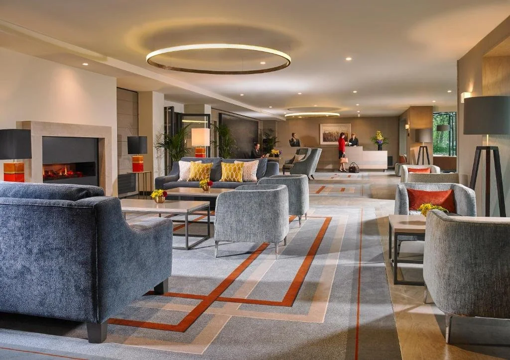 Hotel lobby with seating area featuring sofas and armchairs, a fireplace, and a reception desk with people checking in.
