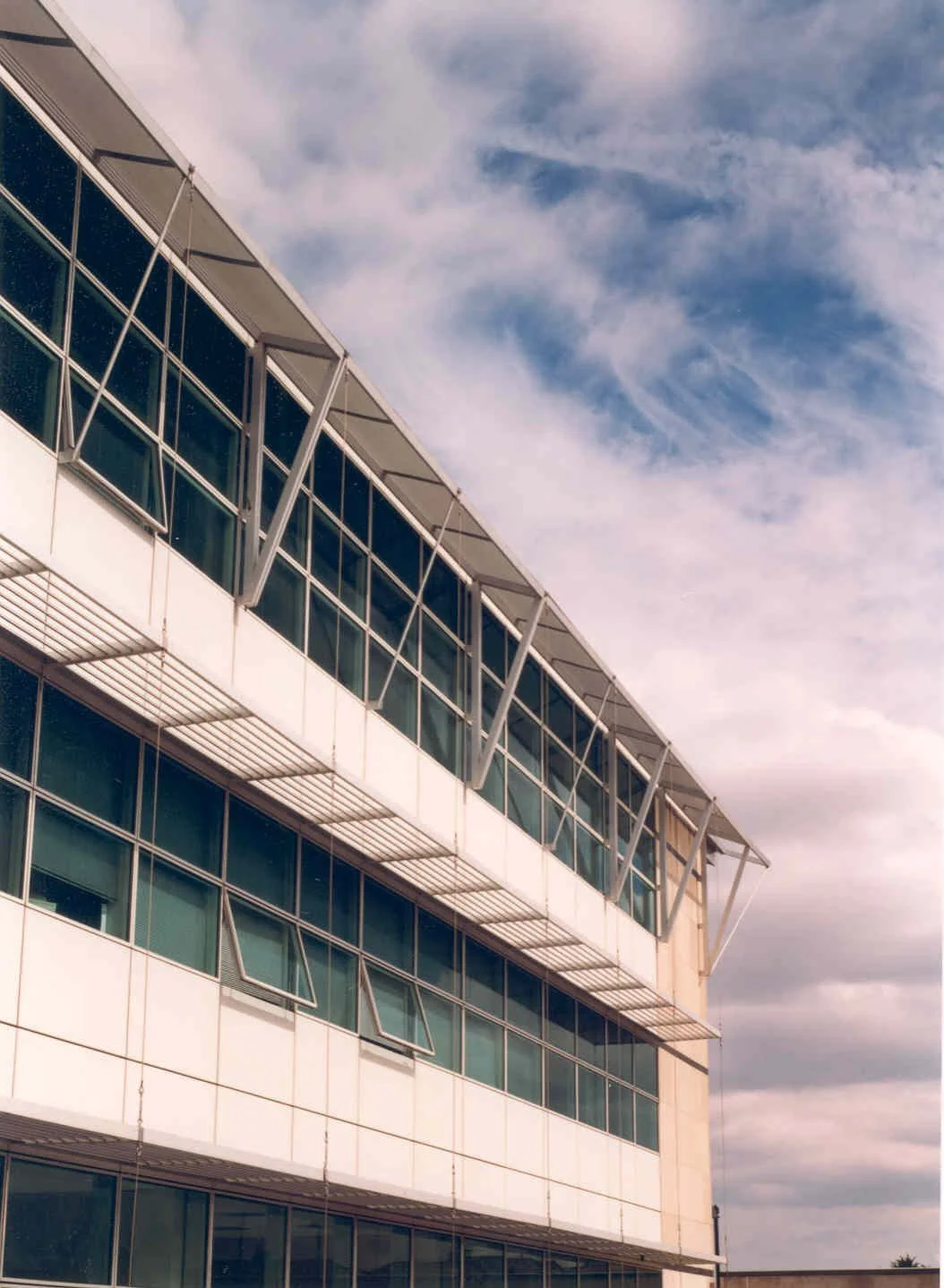 Modern multi-story building with inclined sunshades and glass windows, under a partly cloudy sky.