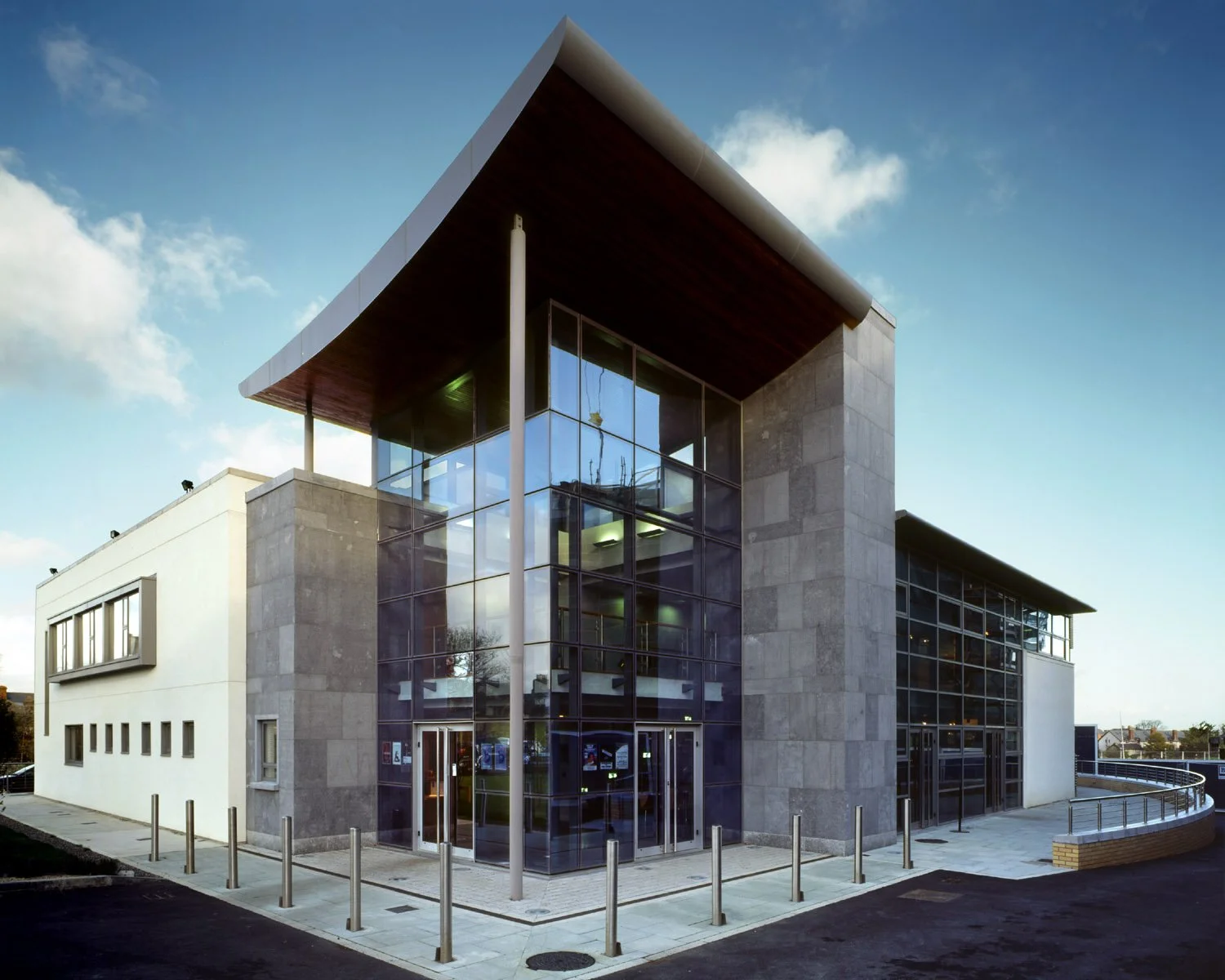 Modern building with large glass windows and a curved roof, surrounded by parking areas and bollards, under a partly cloudy sky.