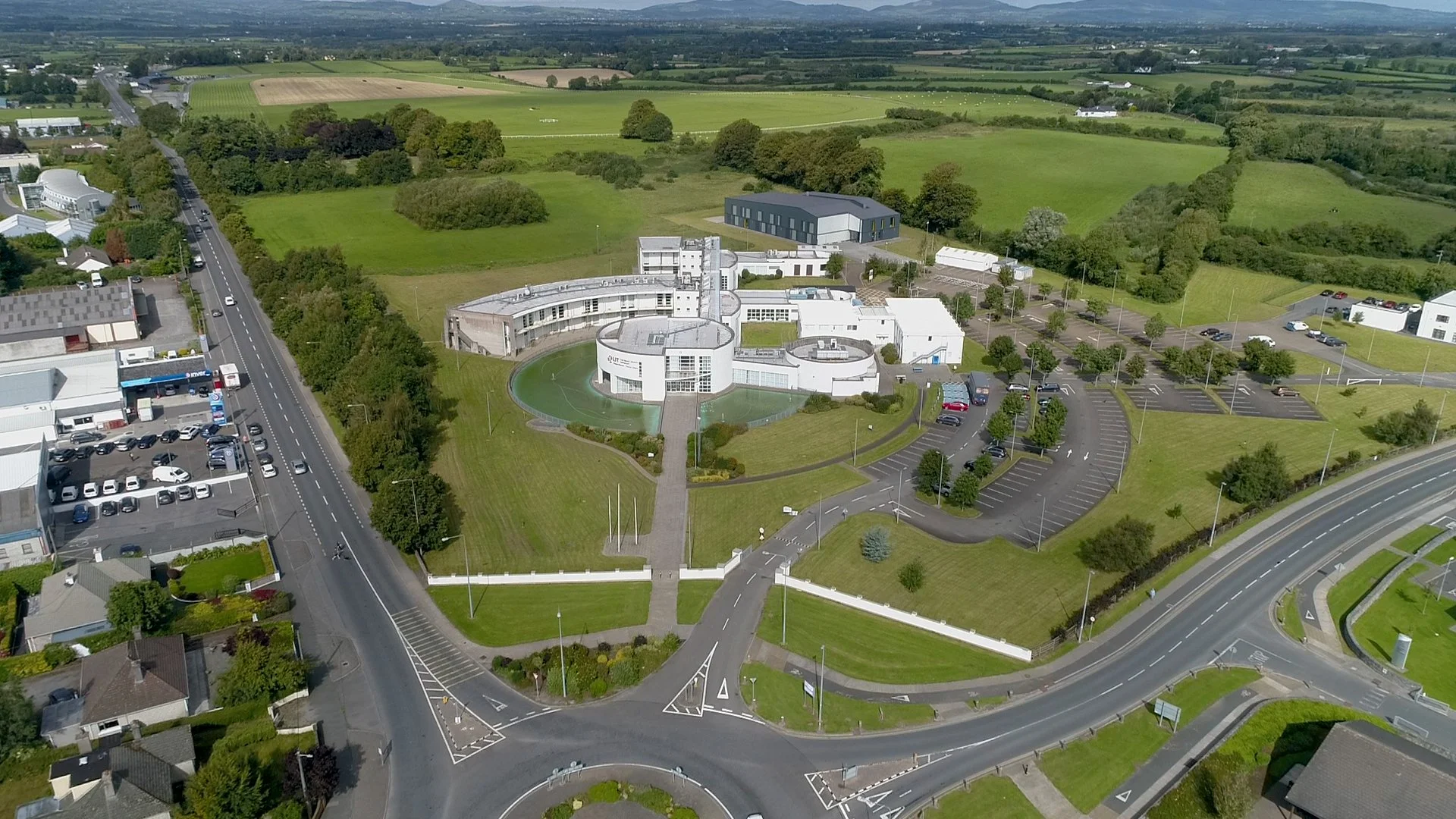 Aerial view of a modern building complex with curved design, surrounded by parking lots, roads, and green landscape, with open fields and trees in the background.