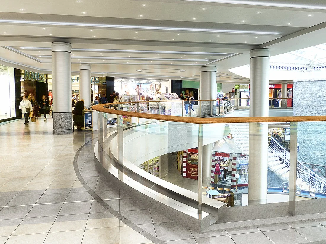 Interior of a shopping mall with multiple stores, shoppers walking, and a glass railing overlooking a lower level with a staircase and storefronts.