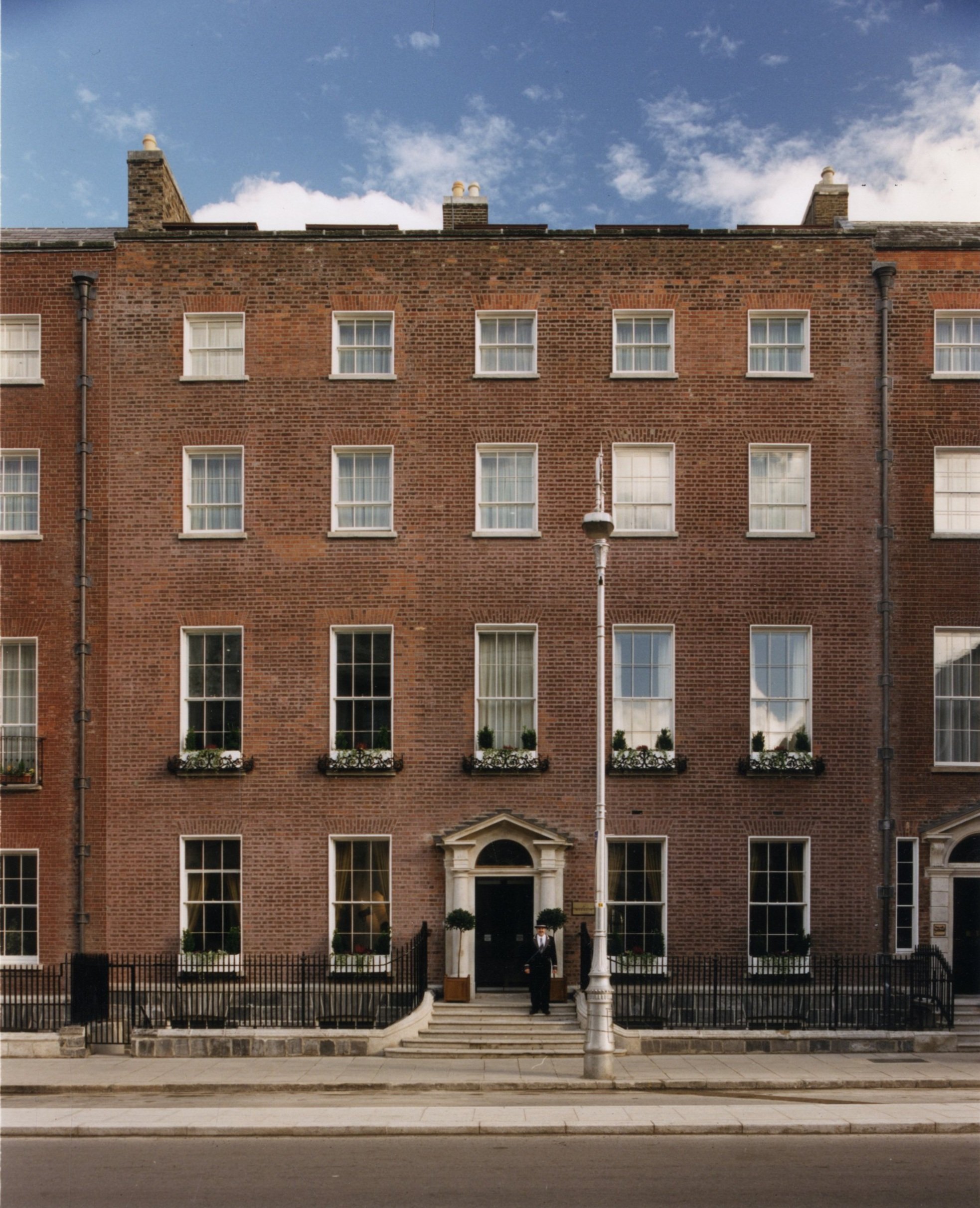 A multi-story brick building with several windows decorated with flower boxes, a central black door, and a person standing at the entrance, with a streetlamp and sidewalk in the foreground, under a partly cloudy sky.