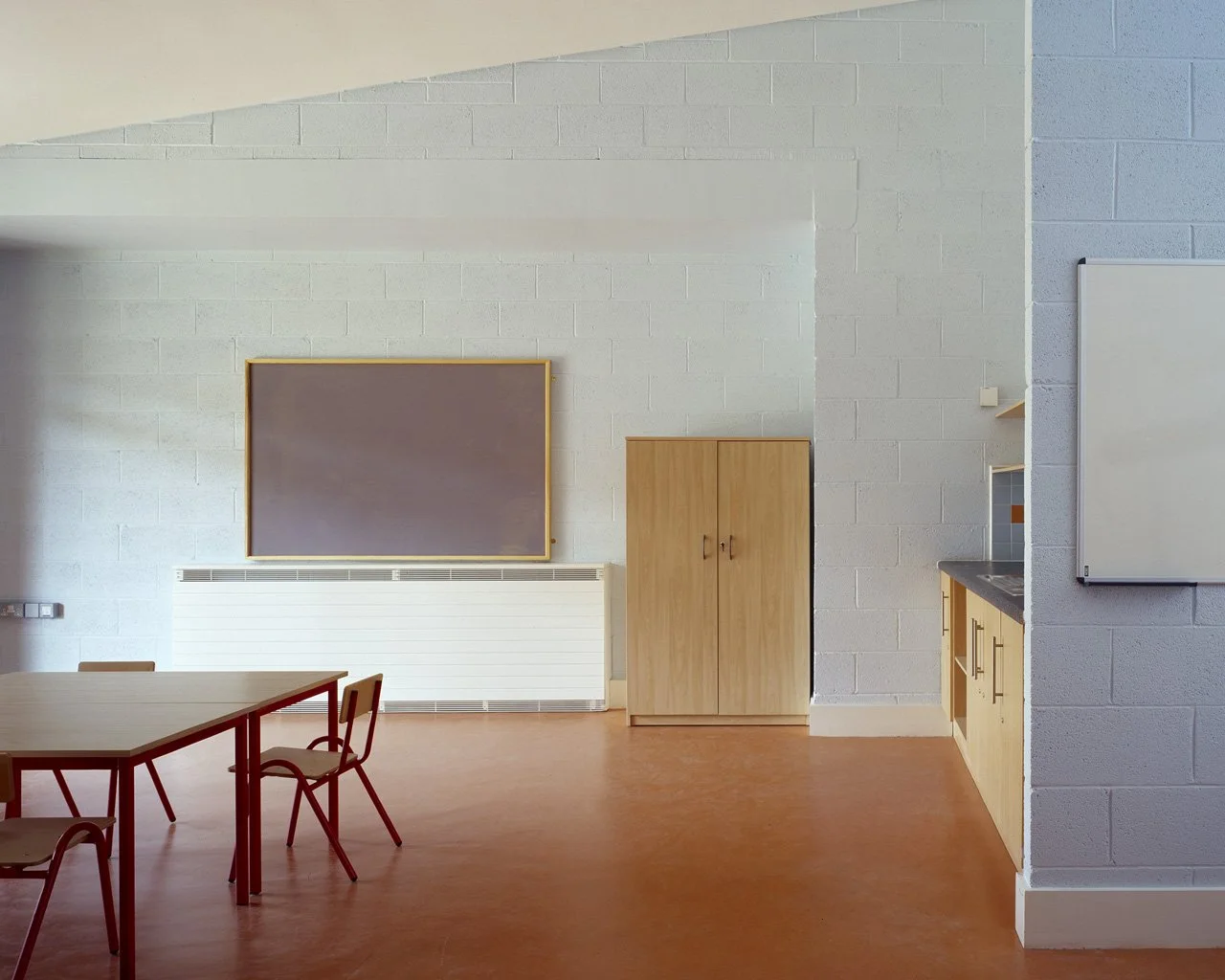 An empty classroom with a white brick wall, a blackboard with a wooden frame, a wooden storage cabinet, a whiteboard, a table with chairs, and a kitchenette area in the background.