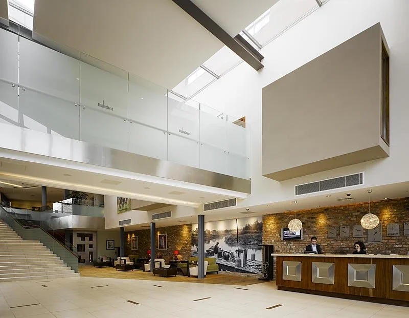 Modern hotel lobby with a brick accent wall, a reception desk with two staff members, seating areas with chairs and tables, a staircase, and ample natural lighting from large skylights.
