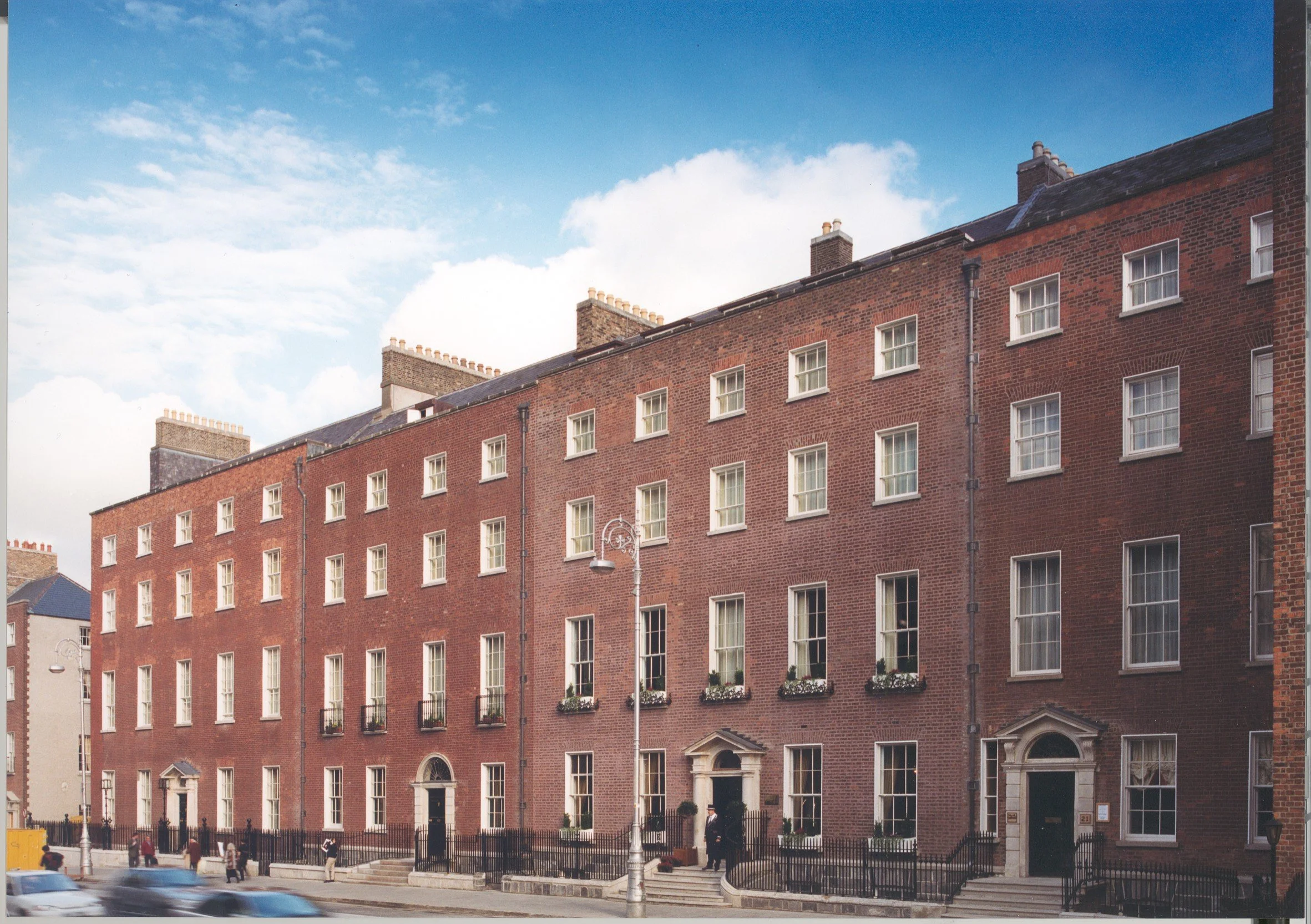 A red brick city apartment building with white-framed windows, some with flower boxes, on a corner street with a black fence and street lamps, under a partly cloudy sky.