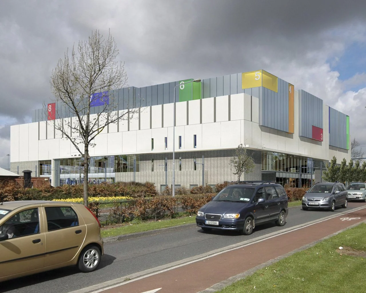 A modern multi-story building with numbered colored sections on the upper facade, parked cars along the street, a tree, and cloudy sky.