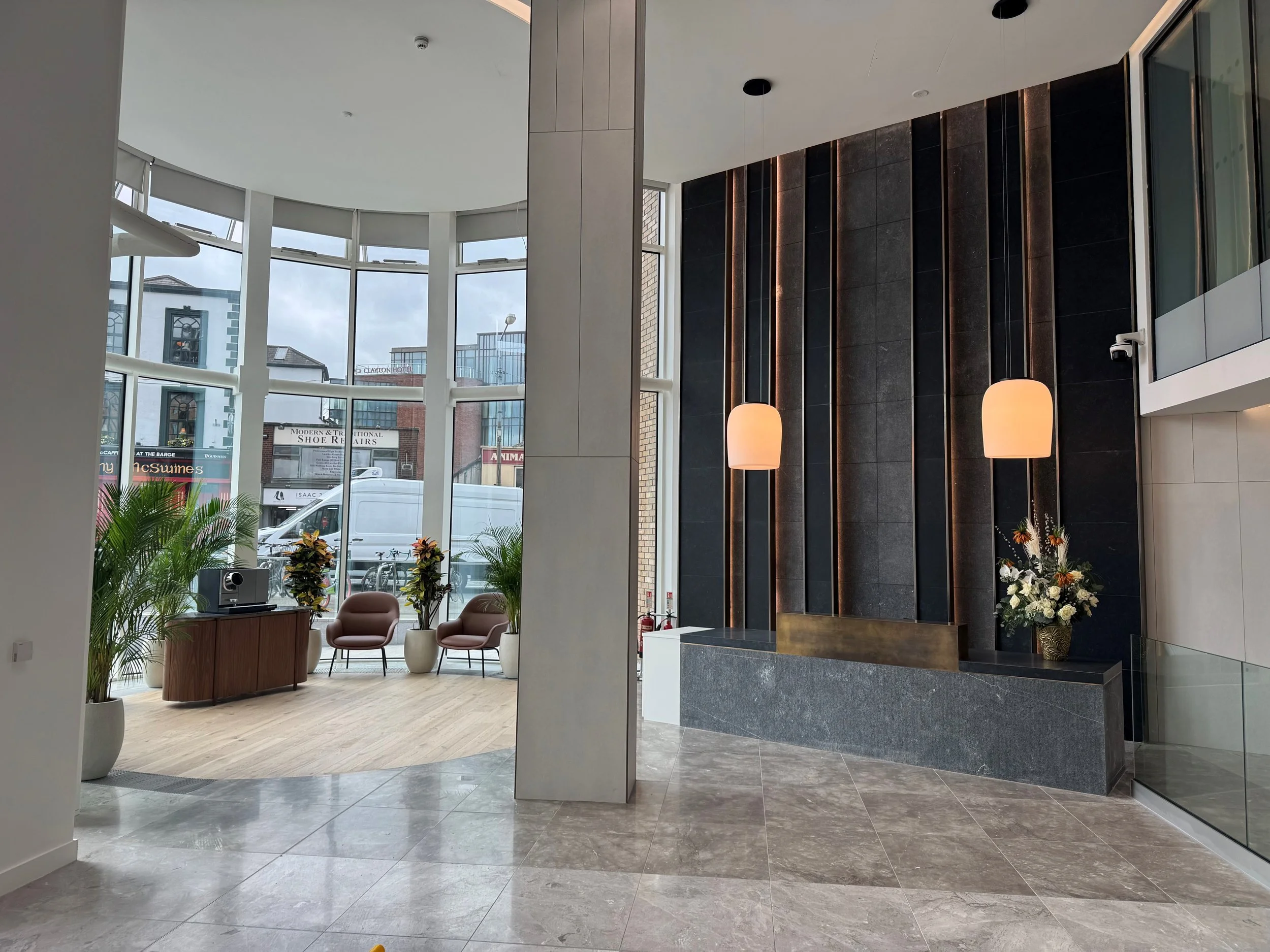Modern hotel lobby with large curved glass windows, seating area with two pink chairs, tall green potted plants, and a reception desk with flower arrangement and wall-mounted lamps with pink shades.