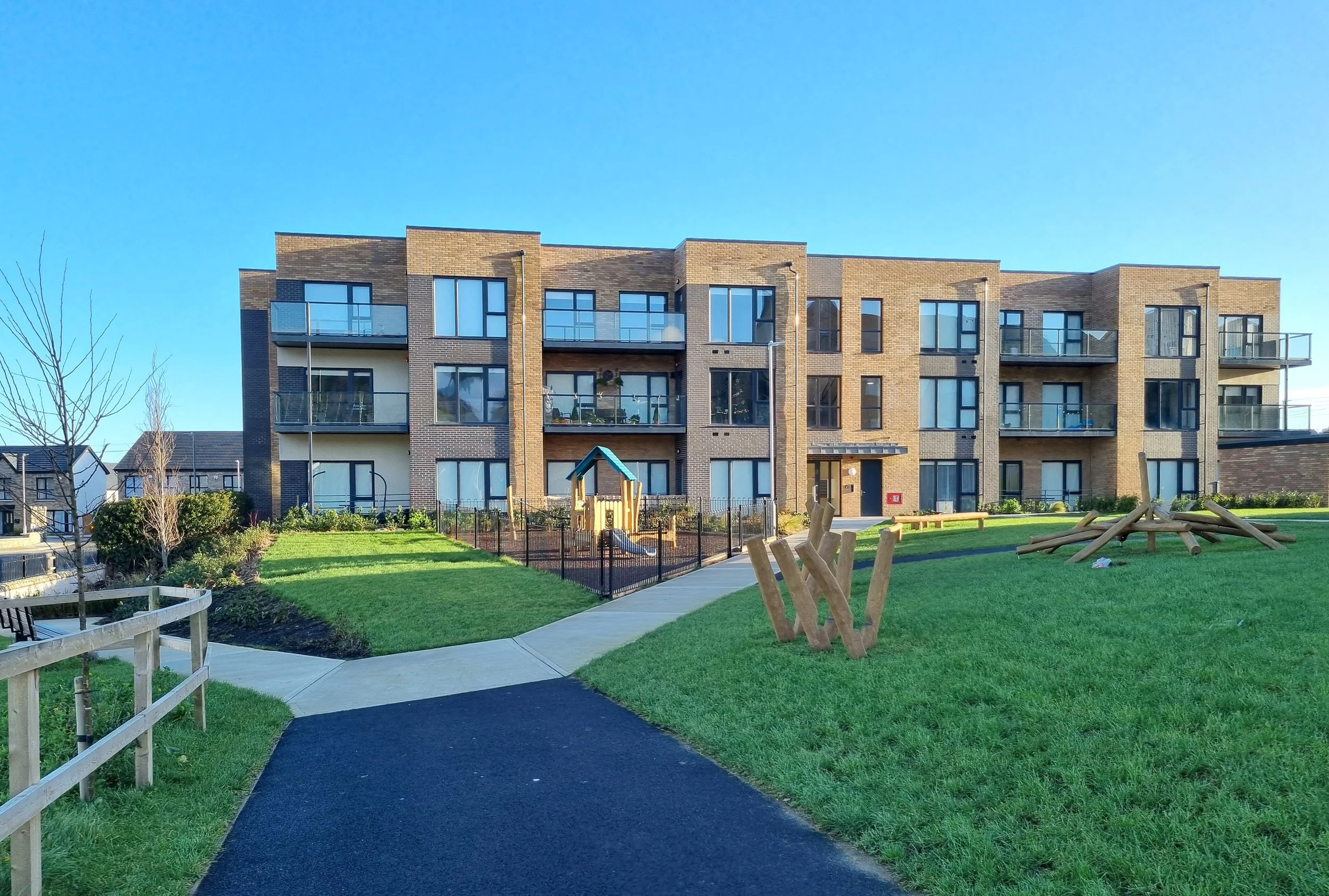 A modern, multi-story apartment building with balconies, surrounded by a well-maintained grassy lawn, walking paths, and outdoor playground equipment on a sunny day.