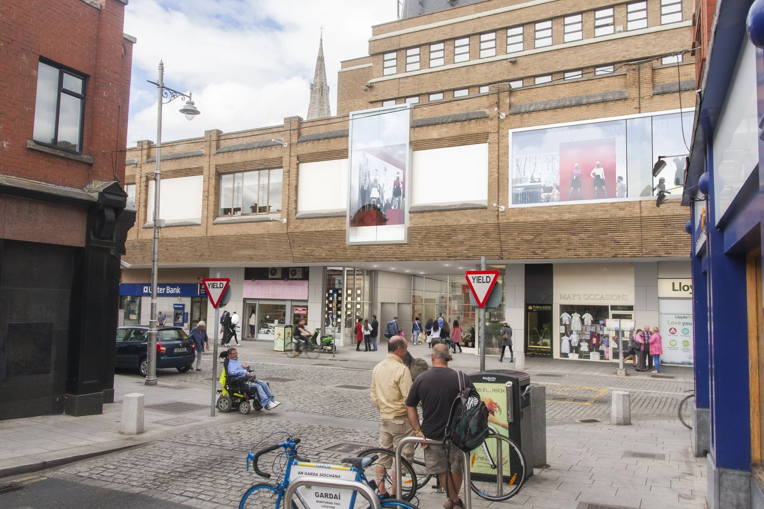 City street scene with pedestrians, cyclists, storefronts, and street signs. There are two men standing near bicycles, a person in a wheelchair, and people walking on the sidewalk. Buildings and storefronts, including a bank and clothing shops, are in the background.
