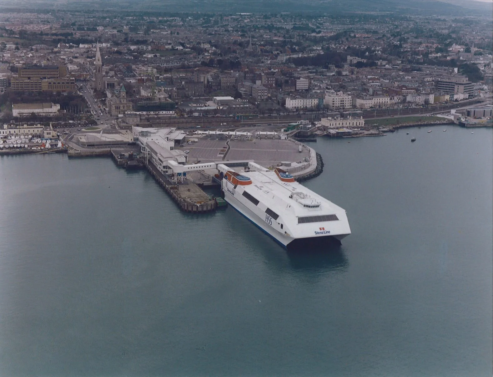 A large ferry boat docked at a pier in a harbor city, with a cityscape and buildings in the background.
