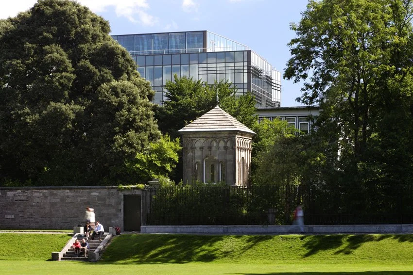 A park with lush green grass and large trees, a small historic stone structure with a peaked roof, a stone wall, and people sitting on steps. A modern glass building is visible behind the trees.