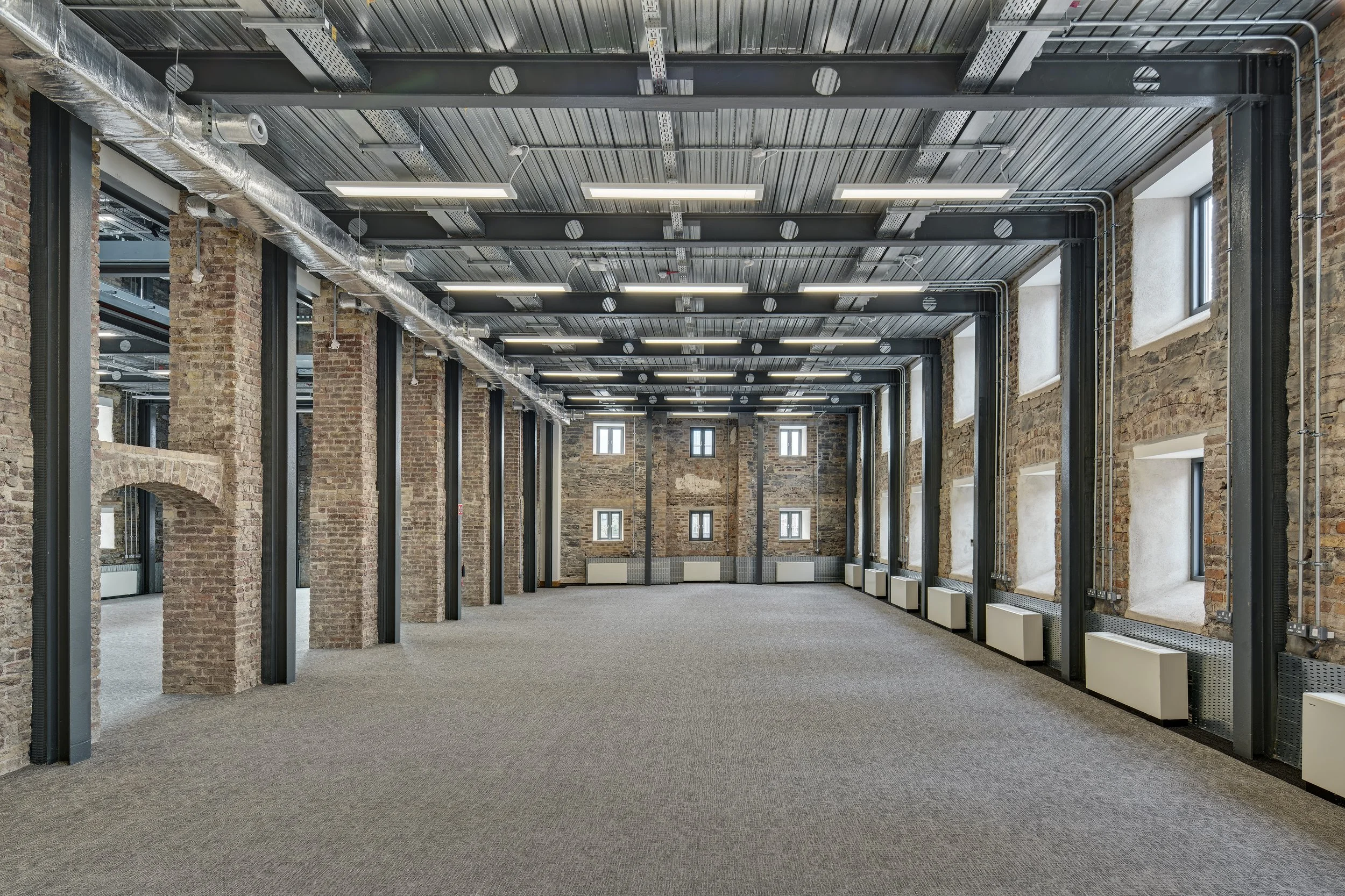 Empty industrial-style office space with exposed brick walls, large windows, gray steel beams, and gray carpeted floor.