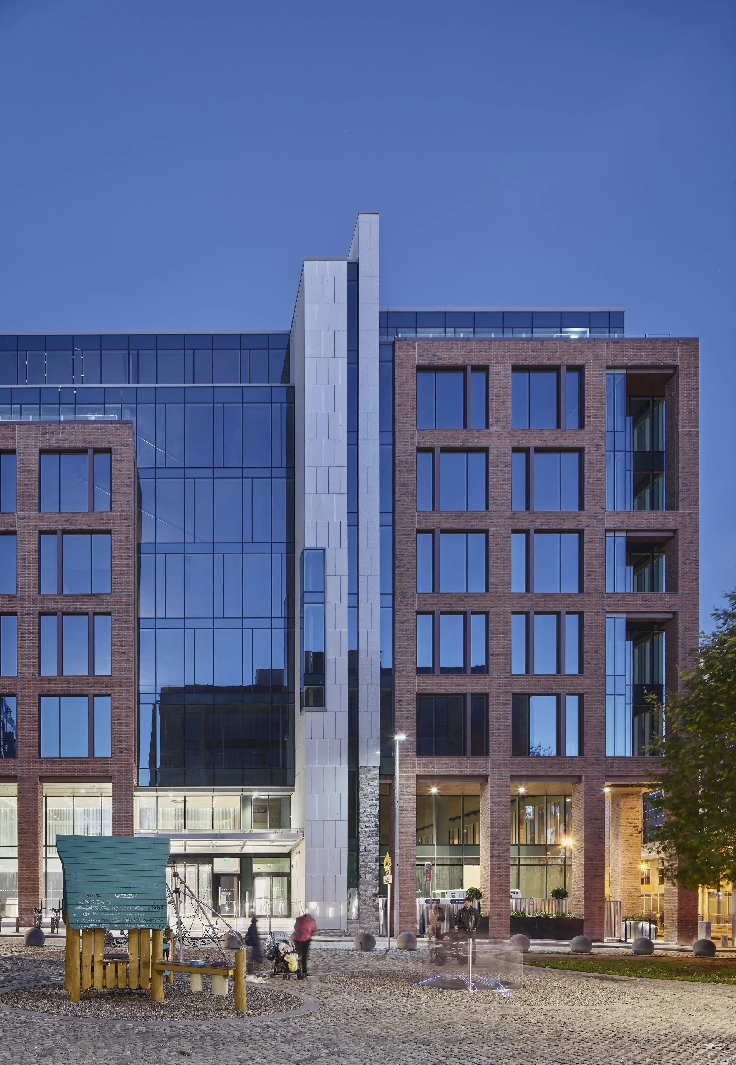 Modern multi-story building with brick and glass exterior, evening sky, people walking in the park in front, with a small fountain and playground.
