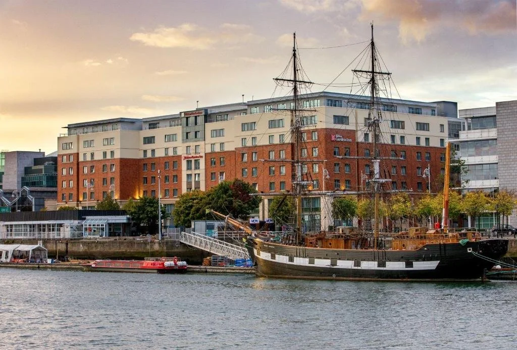 View of a harbor with a historic tall ship docked in front of modern apartment buildings during sunset.
