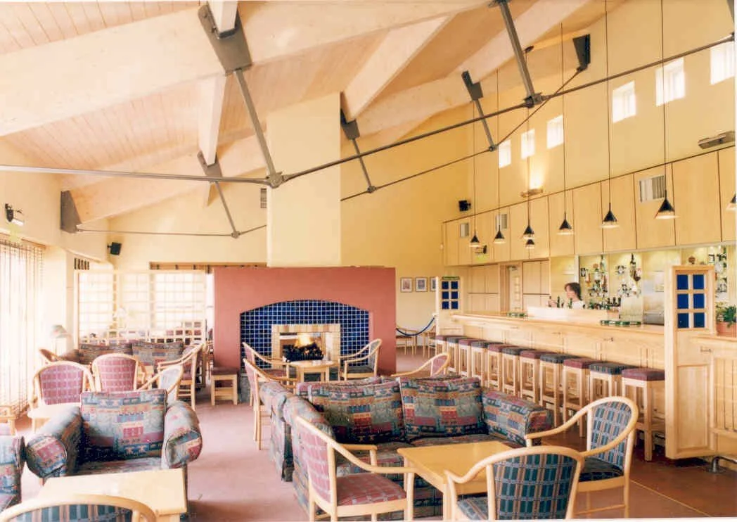 Interior of a cozy lounge or bar area with a fireplace, wooden bar counter with stools, and upholstered seating with patterned fabrics. The room has high ceilings with exposed beams and large windows allowing natural light to fill the space.