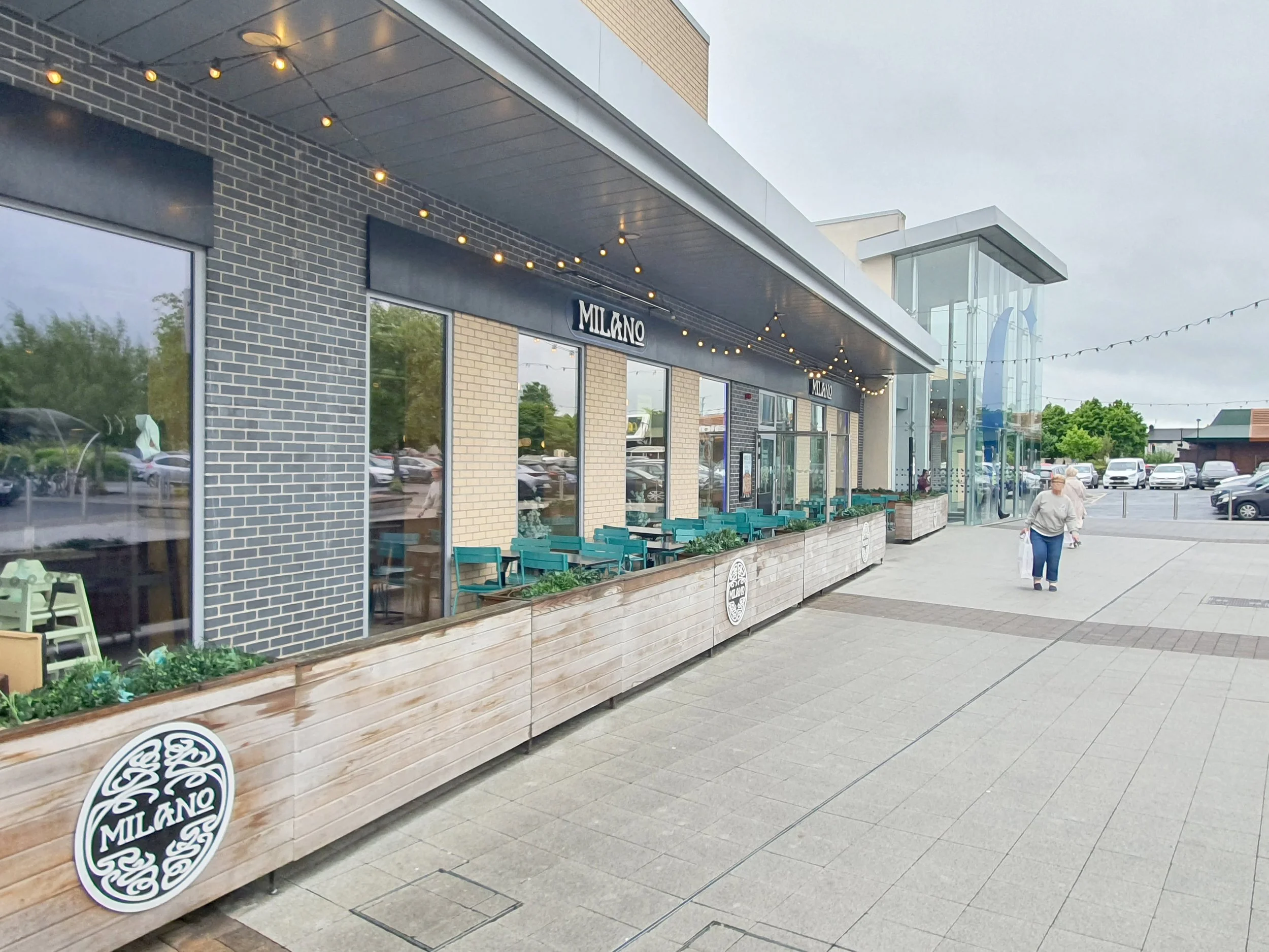 Exterior of a restaurant named Milano with a seating area outside, string lights overhead, and a woman walking on the sidewalk towards the parking lot.