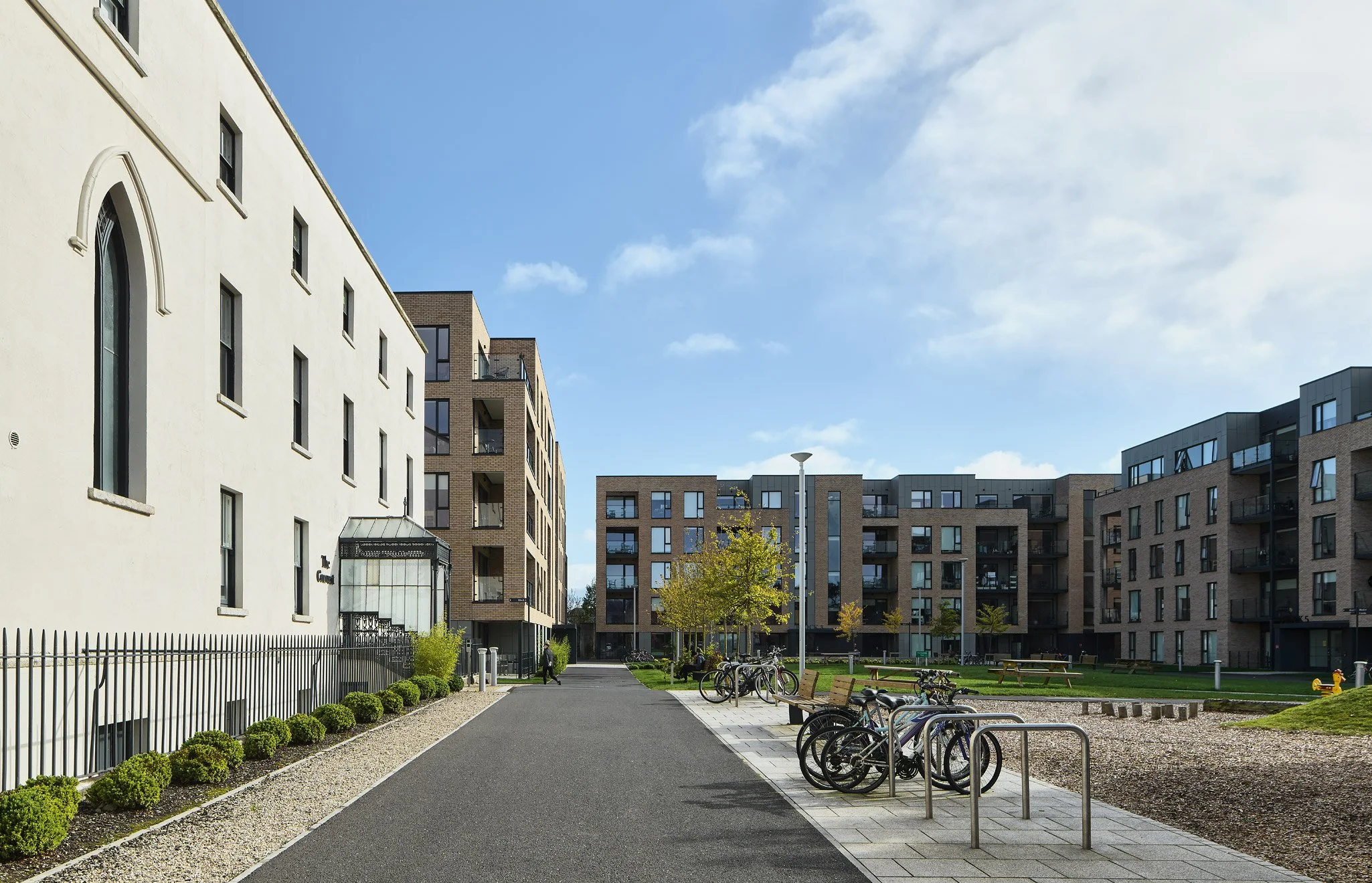 Modern apartment complex with a landscaped courtyard, bike racks, benches, and a paved walkway on a sunny day.