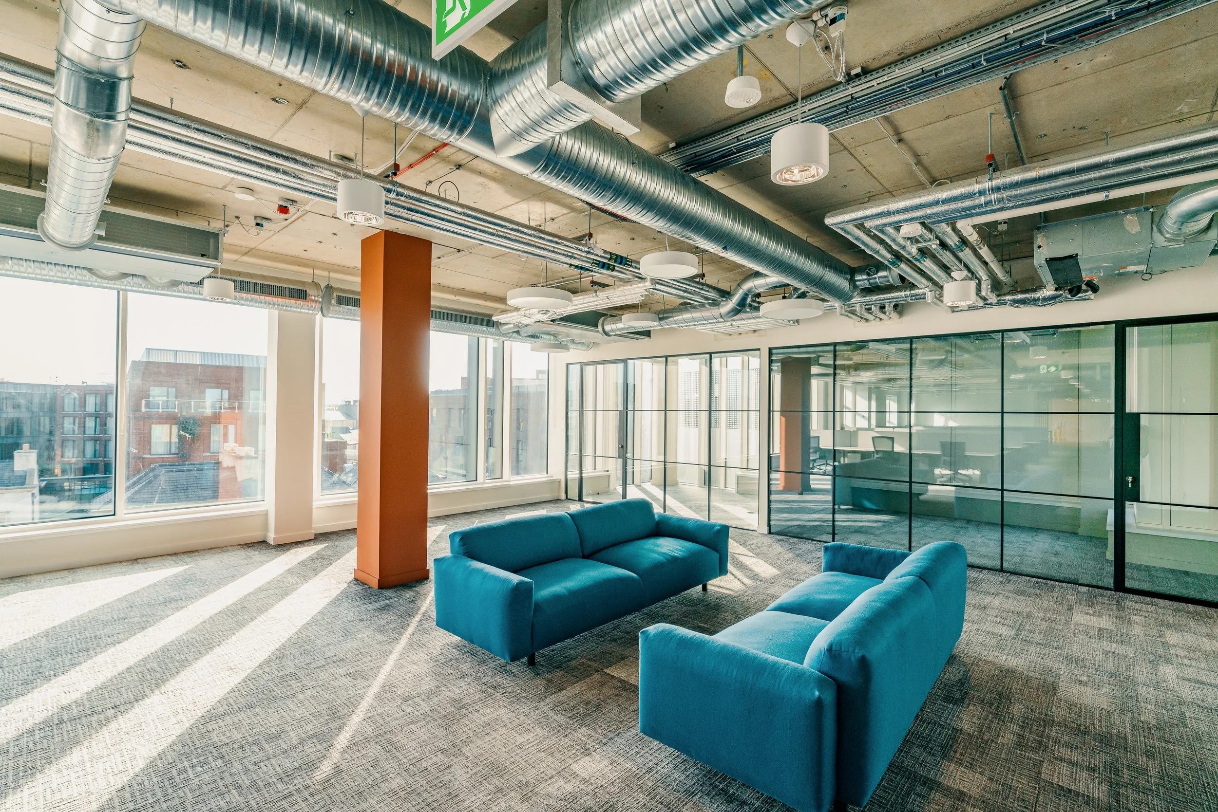 Modern office lounge with two blue sofas, large windows, green glass partition, and industrial ceiling with exposed ductwork.