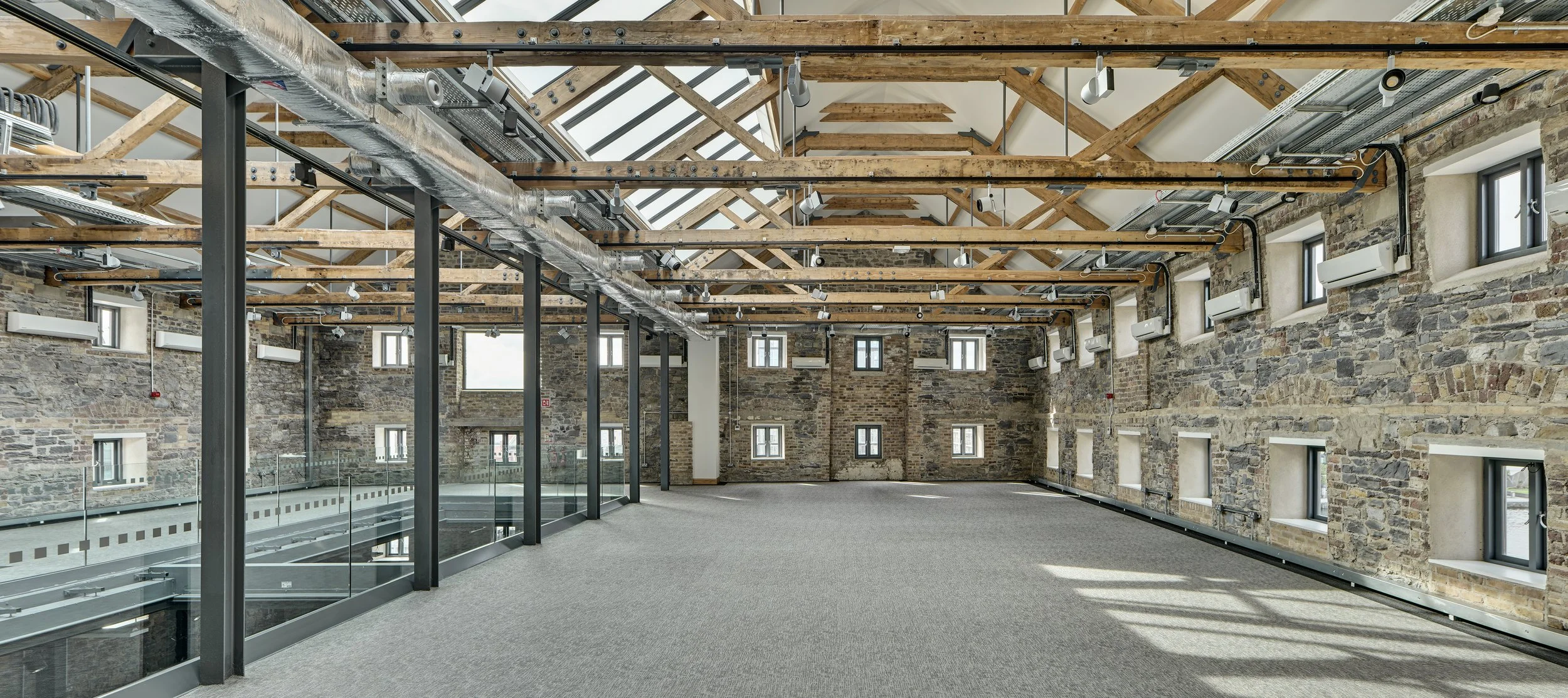 An empty industrial-style loft with exposed brick walls, wooden beams, and skylights. Modern vents and air conditioning units are mounted on the walls, and a glass railing runs along a lower level visible on the left side.