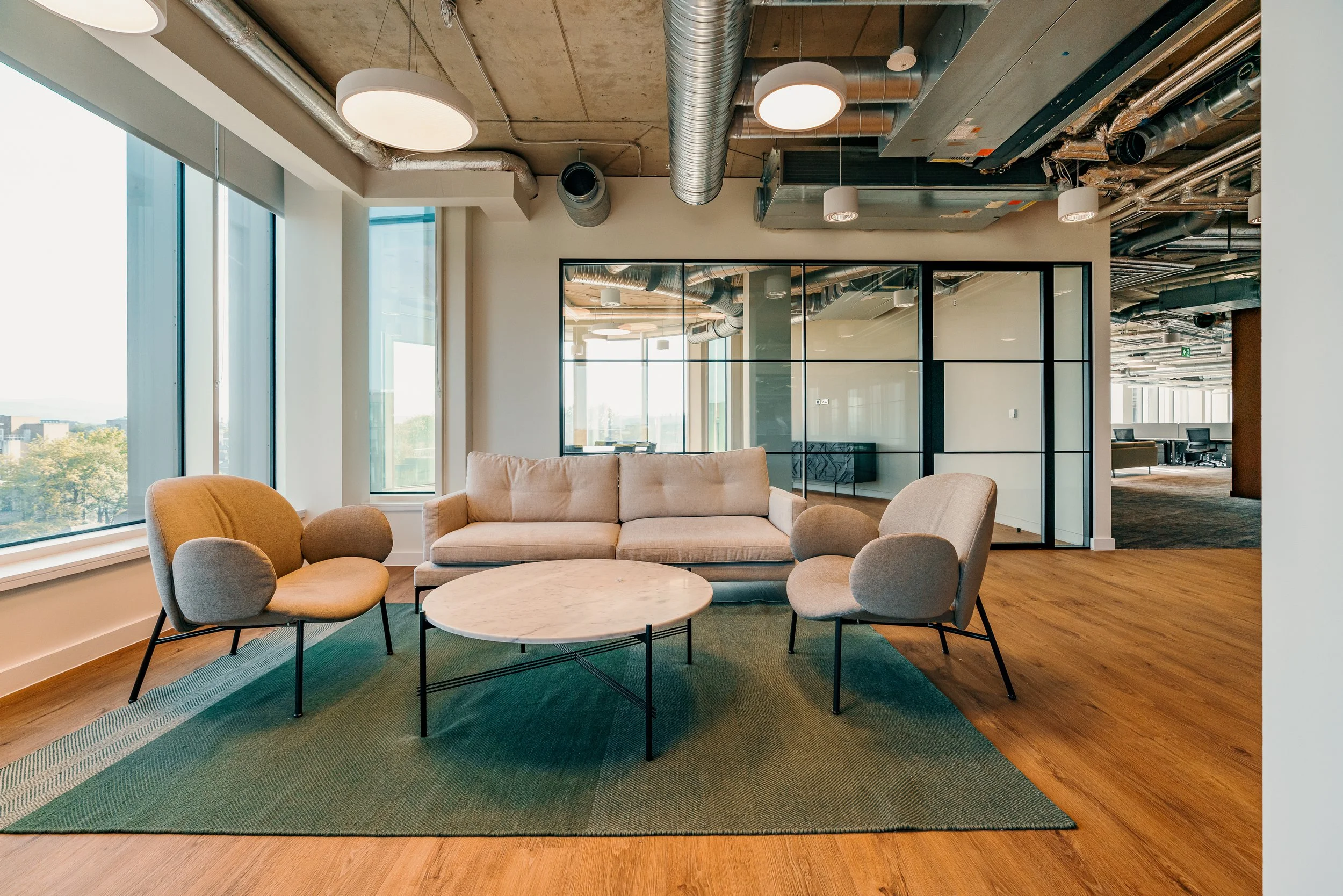 Modern office lounge area with beige sofa, two beige armchairs, a round marble coffee table, large windows, exposed ceiling ducts, and glass partitions.