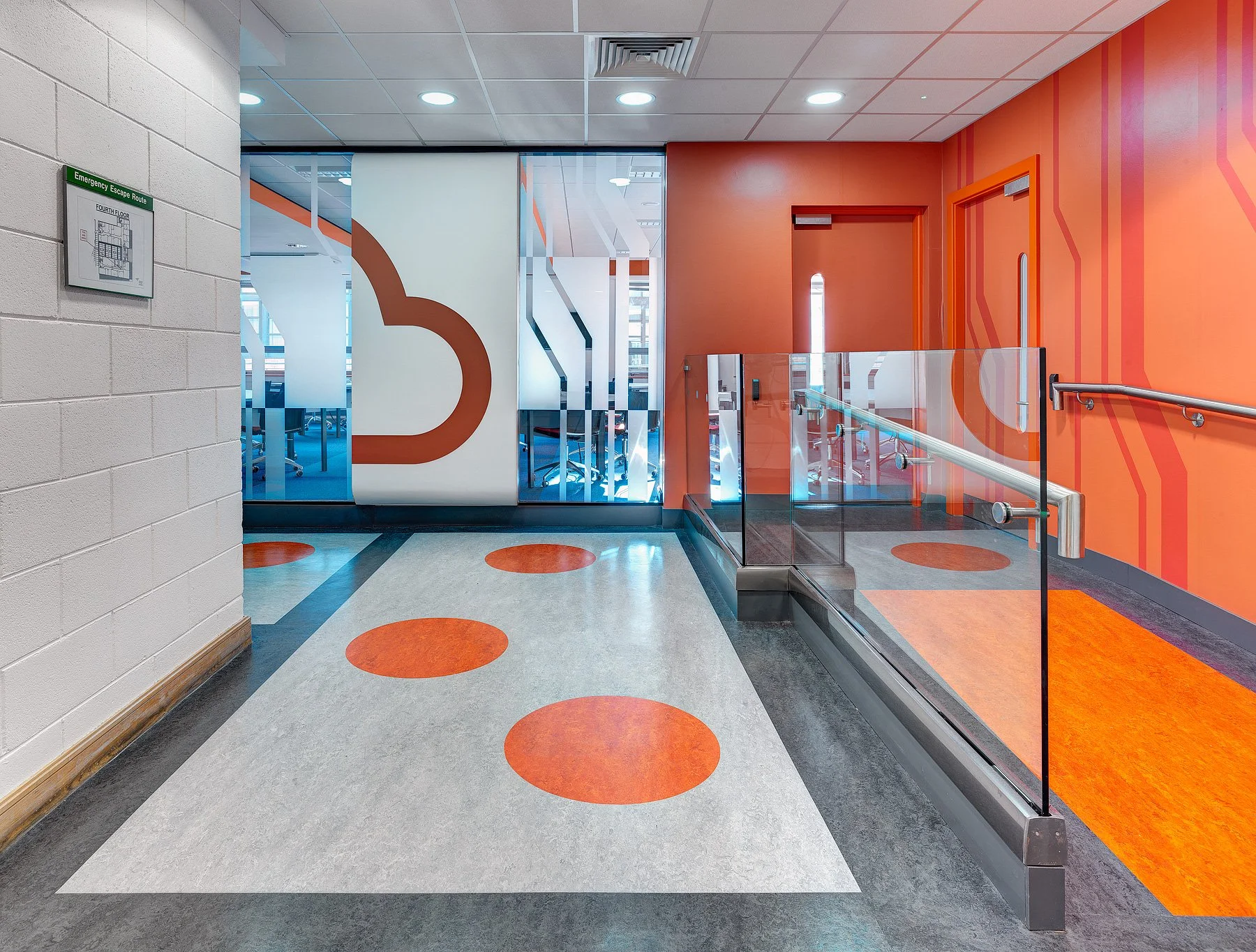 Office hallway with orange and white walls, glass doors with frosted designs, orange-colored flooring, and a gray and white floor with orange circular patterns.