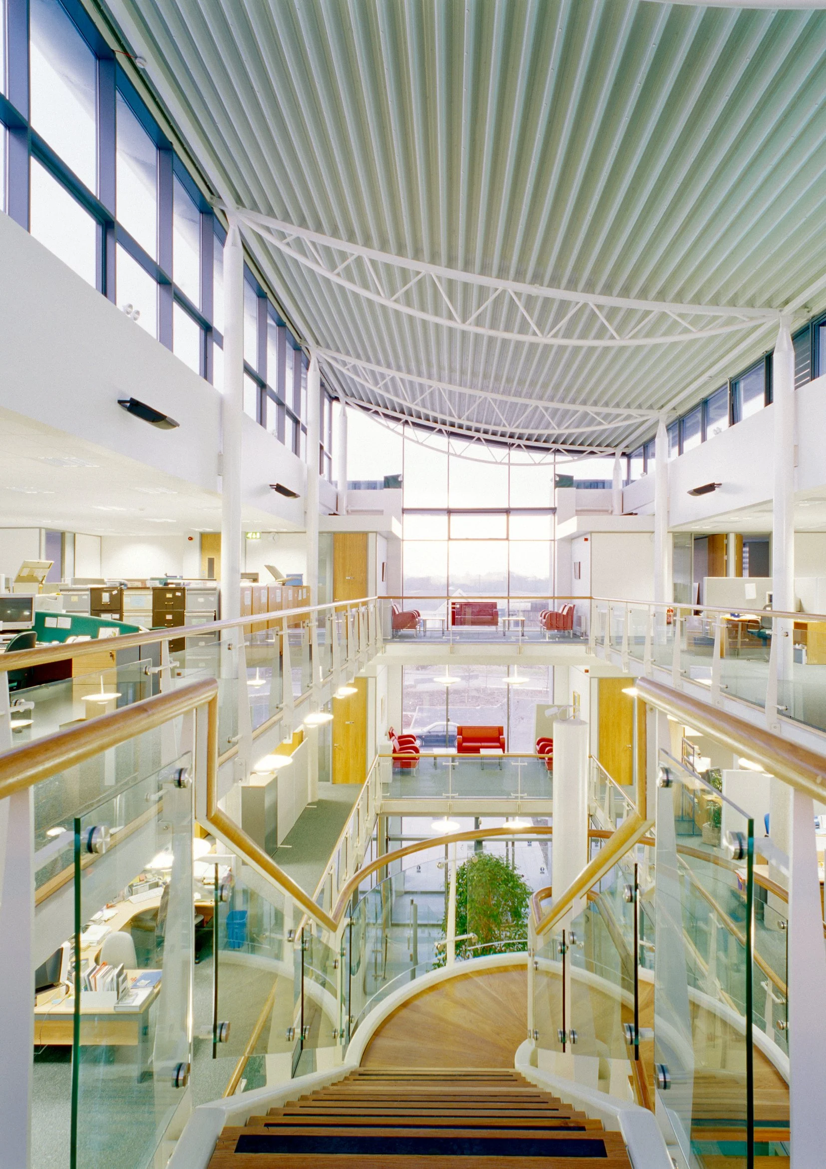Interior of a modern multi-story office building with a glass-walled staircase, glass railings, and sunny seating areas on different floors.