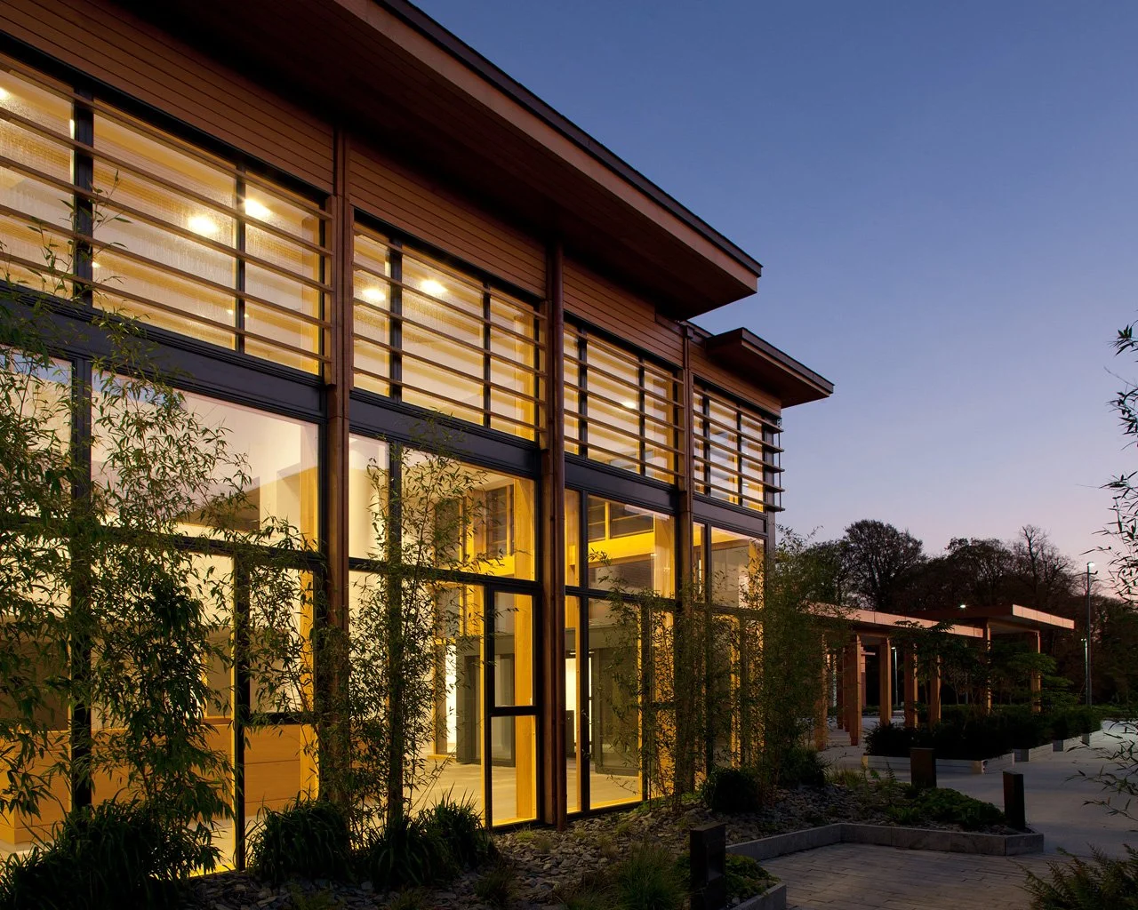 Modern building with large glass windows illuminated from inside, surrounded by landscaped greenery at dusk.