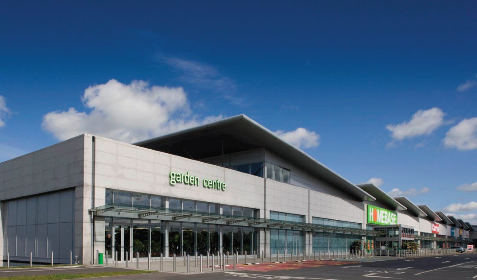 Modern shopping center with signs for Garden Centre, Homebase, and other stores, under a blue sky with clouds.