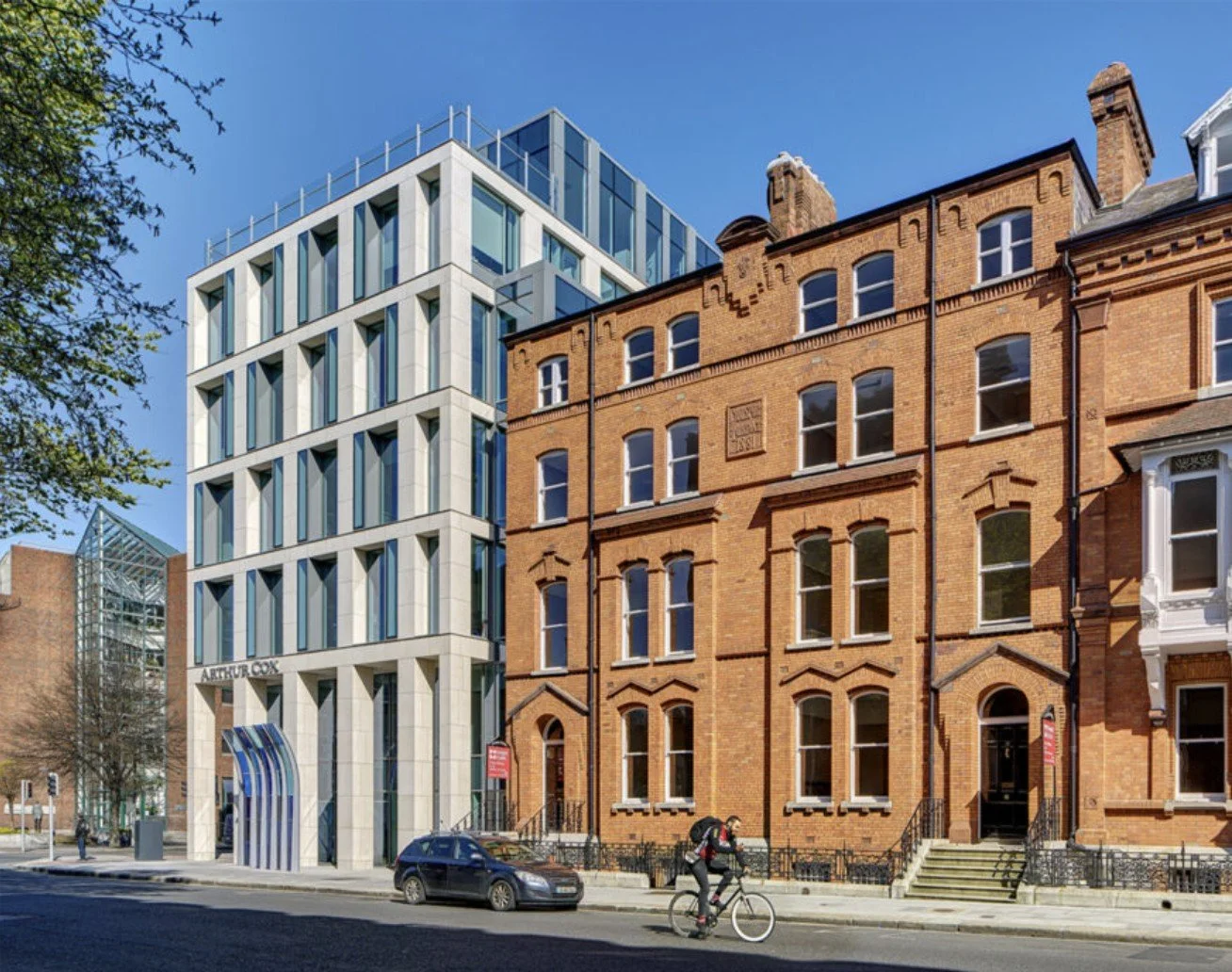 Street view with a modern white building and older brick buildings, cyclist riding on road, parked car, clear blue sky, some trees
