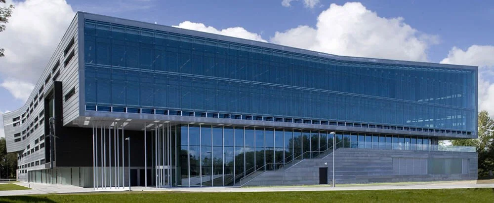 Modern office building with glass exterior and vertical white structural elements, set against a partly cloudy sky.