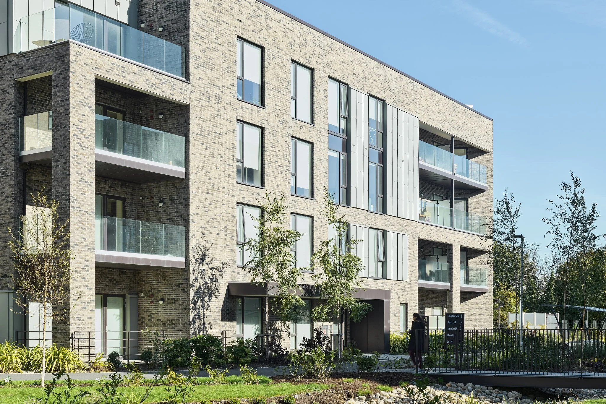 Modern multi-story apartment building with brick exterior and glass balconies, surrounded by landscaped greenery and trees, under a clear blue sky.