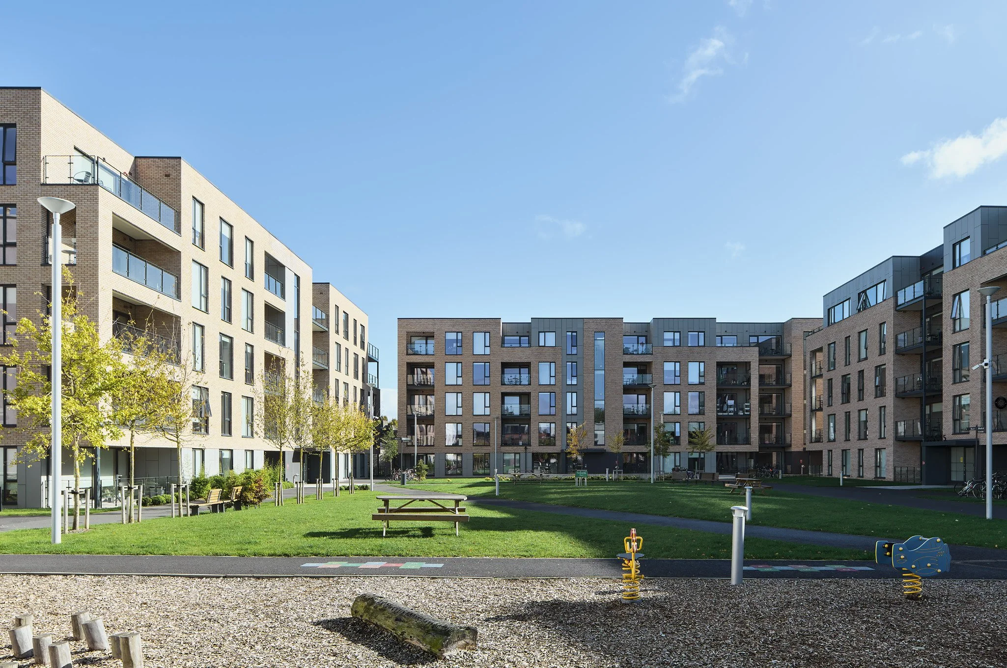 A modern apartment complex with multiple buildings surrounding a green courtyard with benches, trees, playground equipment, and a paved walking path under a clear blue sky.