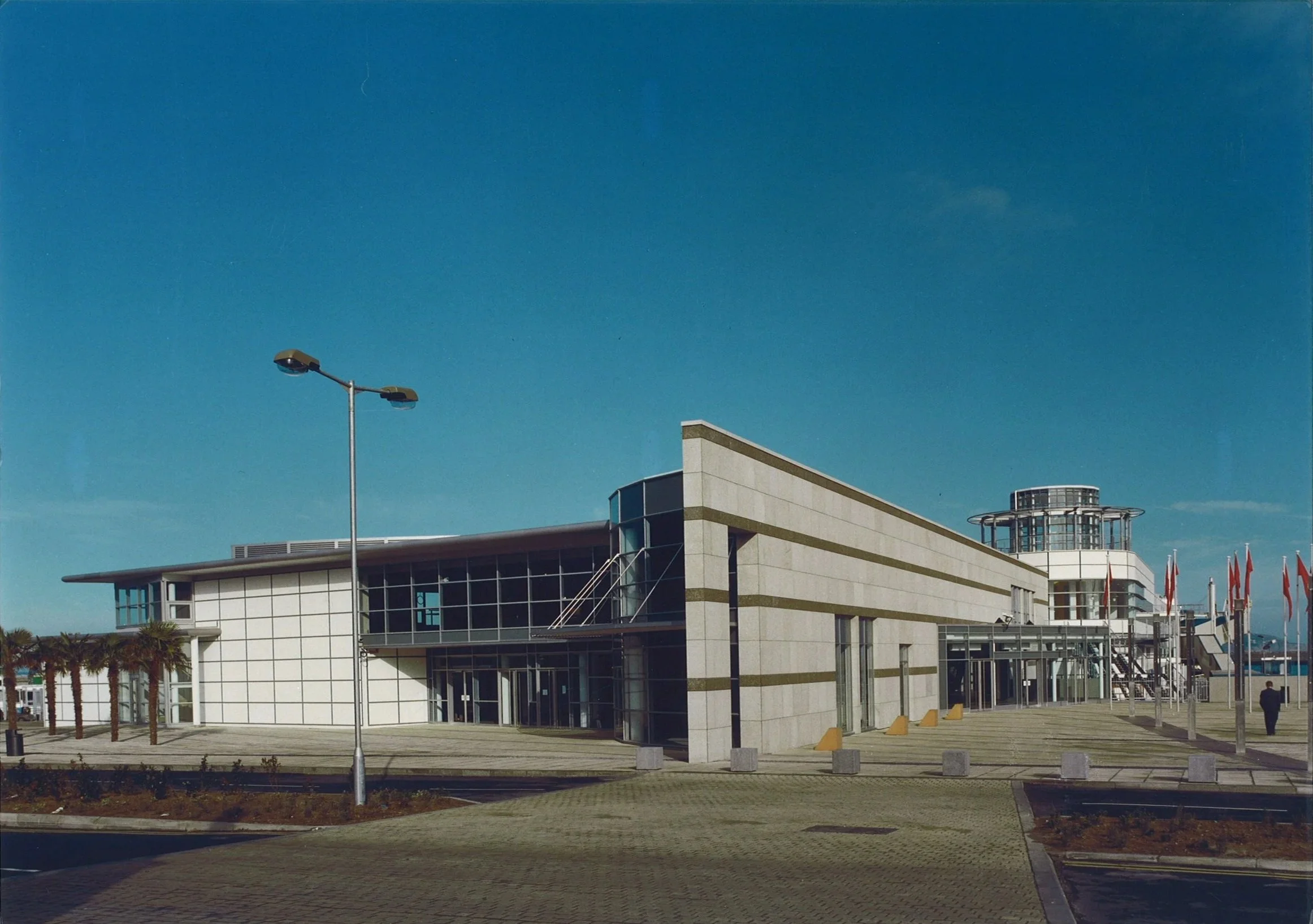 Modern building with glass windows, palm trees to the left, lamppost in front, and a row of flags on the right, under a clear blue sky.