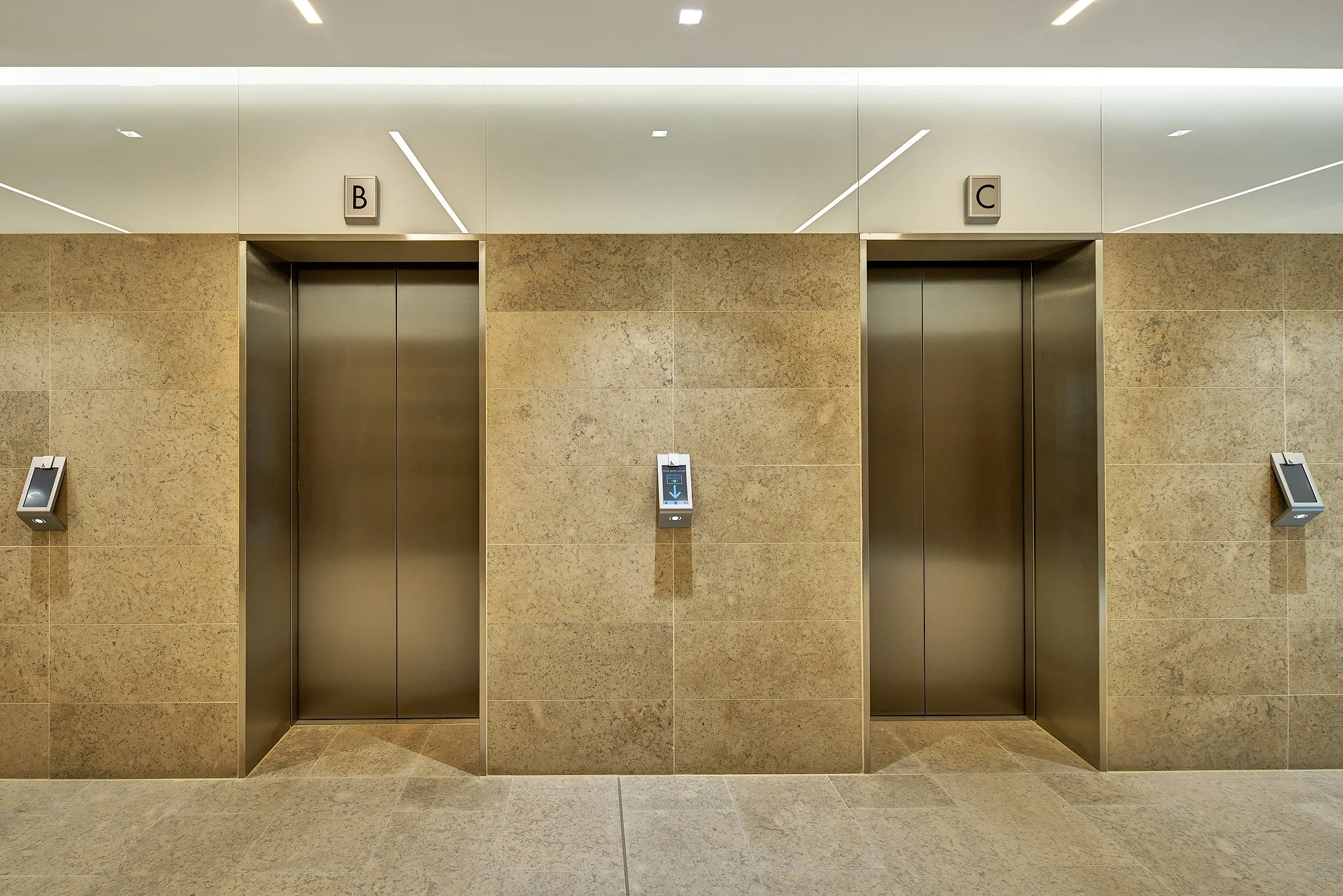 Two elevator doors in a modern building, with signs labeled 'B' and 'C' above each elevator, and a digital floor indicator between them.