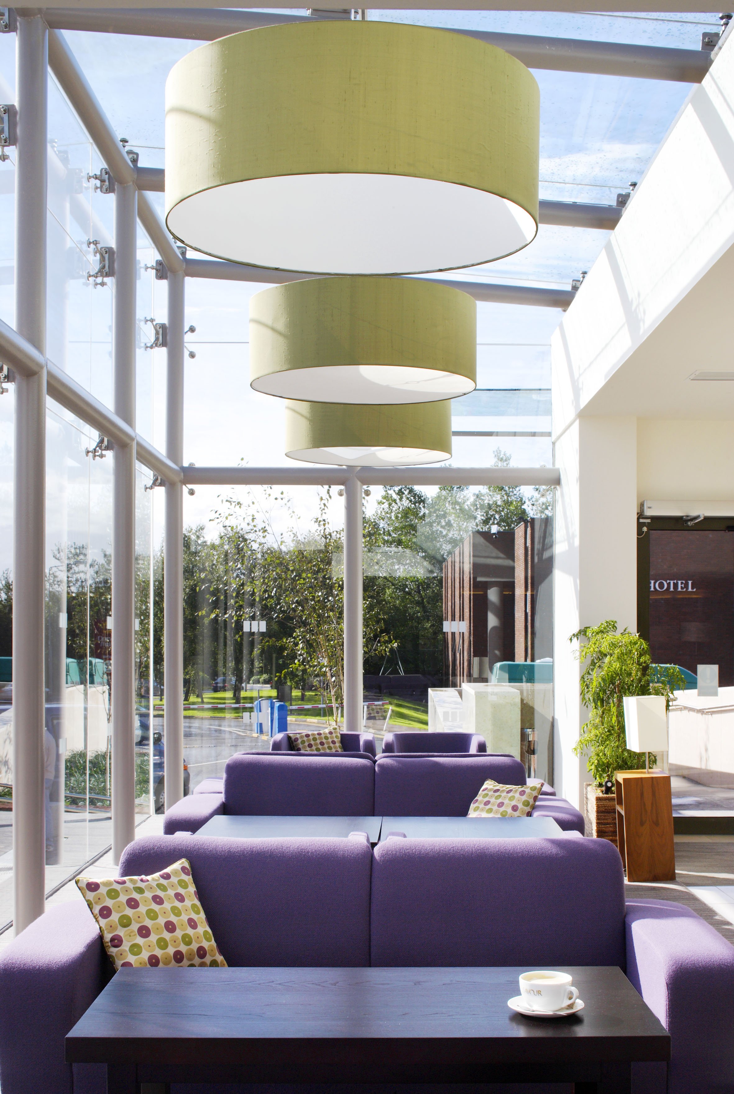 Interior of a hotel lobby with purple sofas, patterned pillows, a table with a cup, and large green pendant lights hanging from a glass ceiling.
