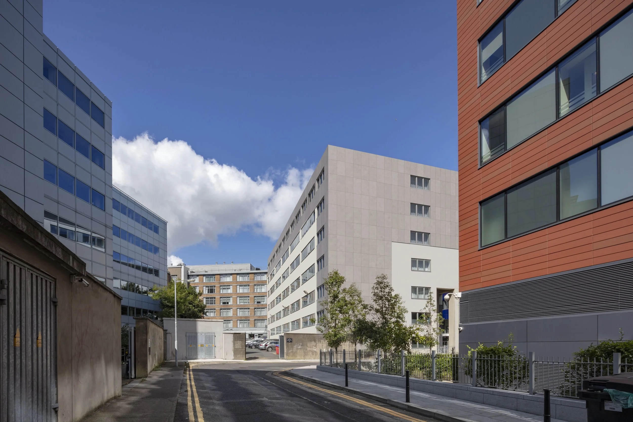 City street lined with modern office buildings under a blue sky with some clouds, and a small green tree in the foreground.
