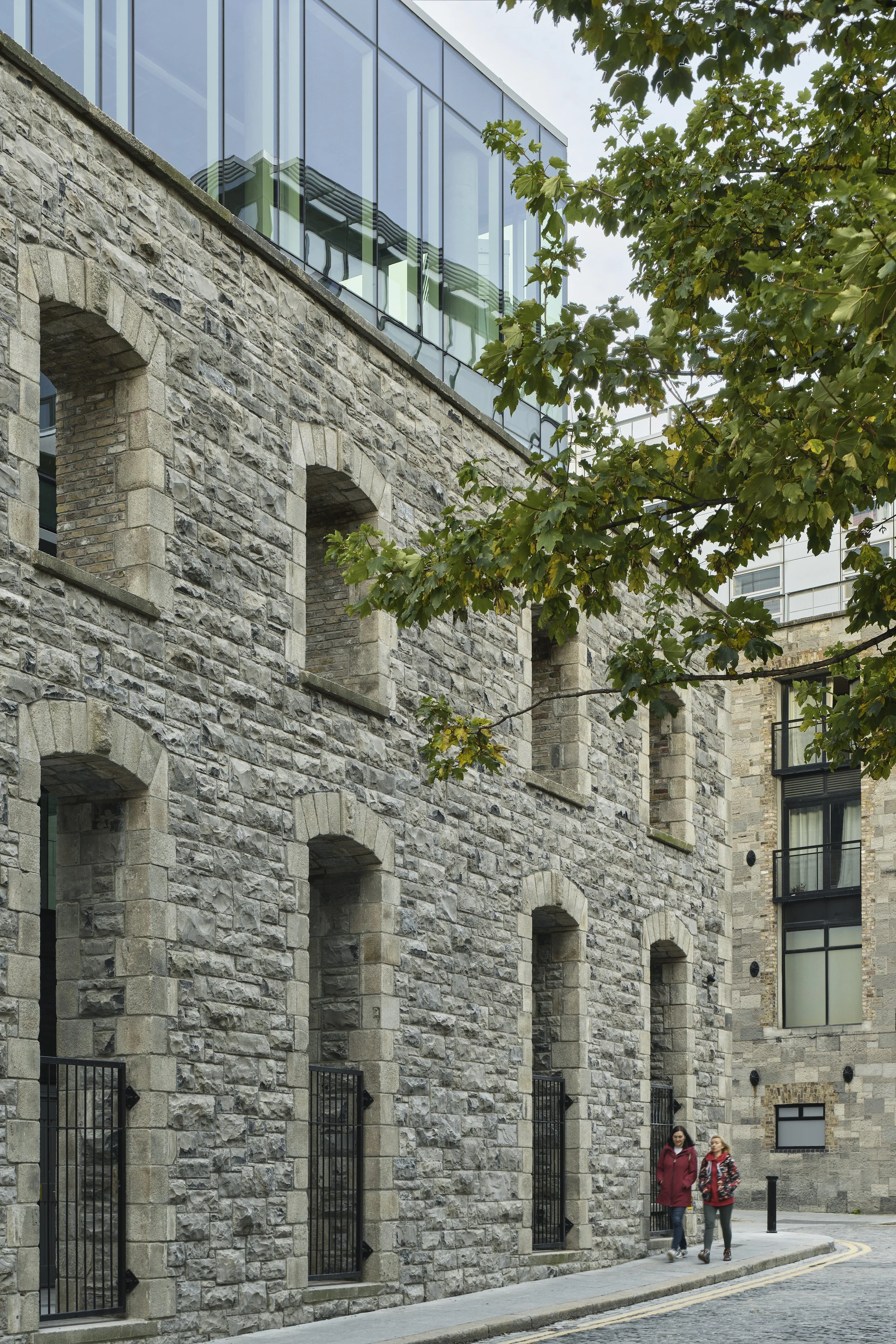 Two women walking along a cobblestone street beside a stone building with arched windows and iron fences, with a tree and modern glass building in the background.