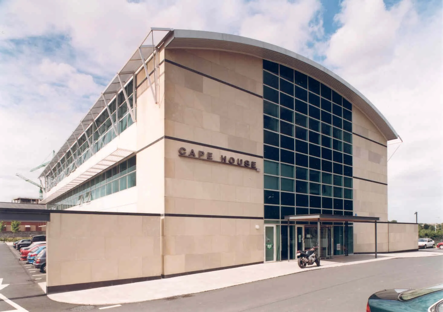 Modern multi-story building with the sign 'Cape House' on the exterior wall, featuring large glass windows, a curved roof, and a parking lot with cars and a motorcycle in front.