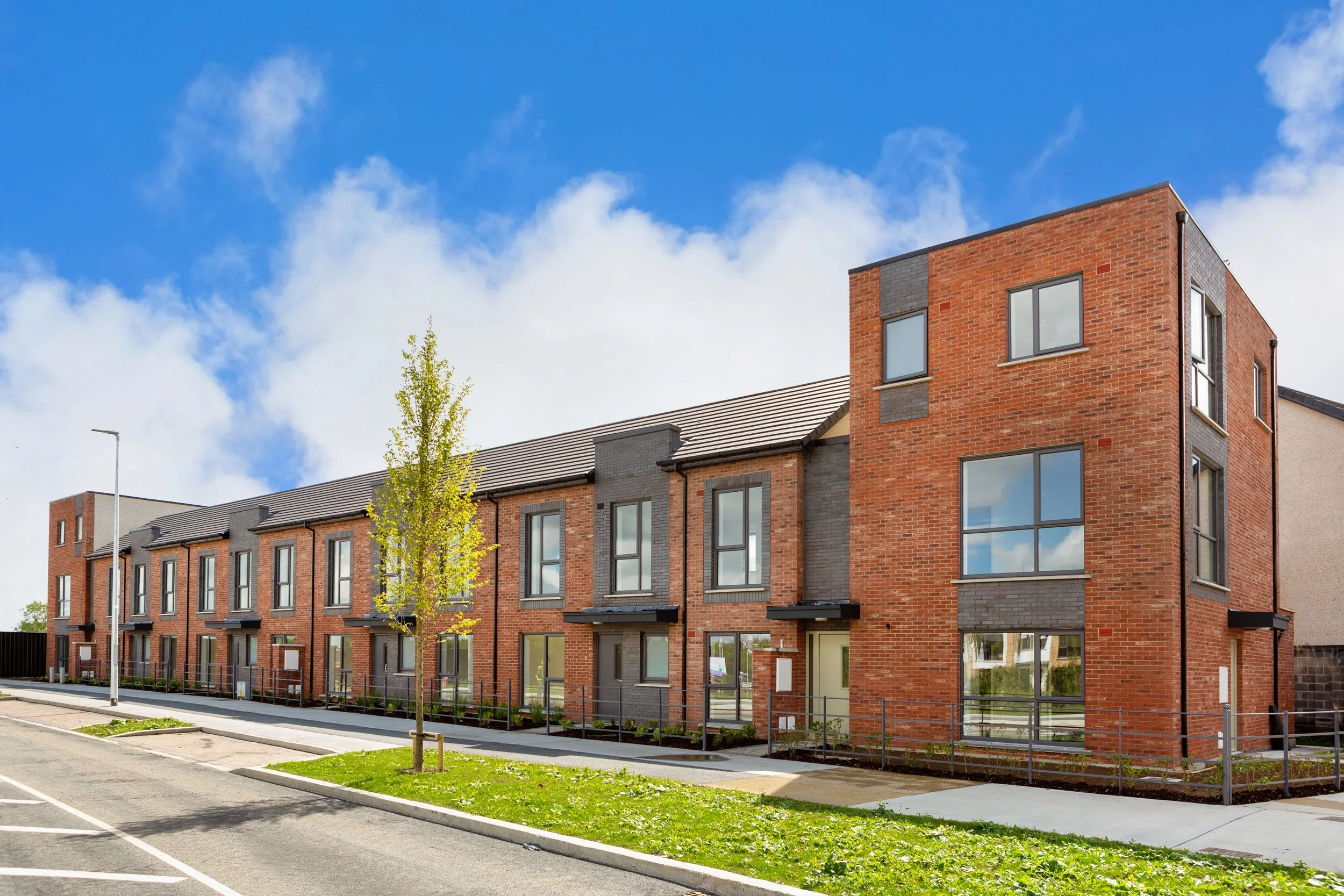 A modern residential apartment complex with brick exterior and large windows, situated along a sidewalk with a small grassy area and a tree, under a partly cloudy blue sky.