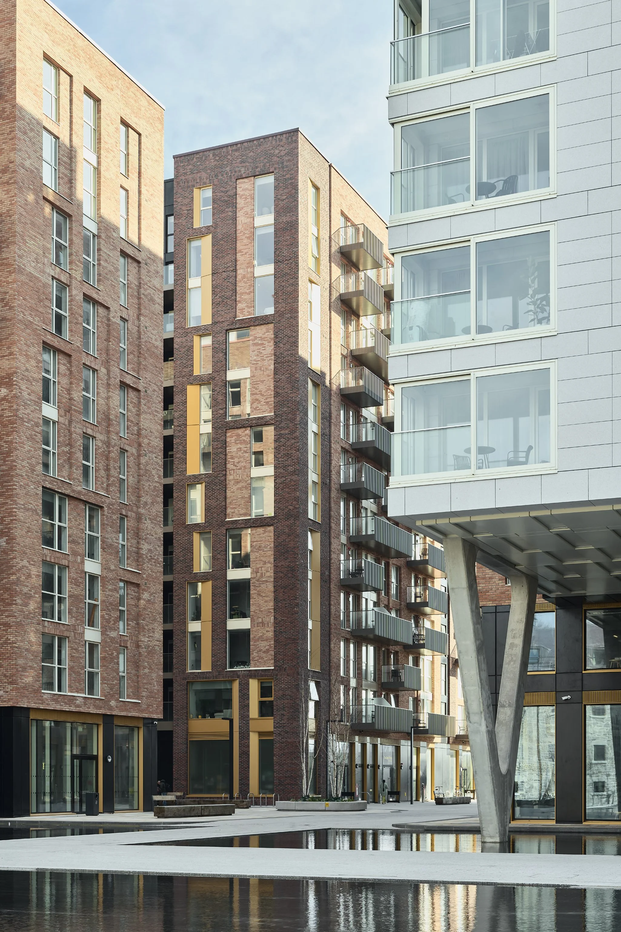 A modern urban courtyard surrounded by multi-story residential buildings with brick and glass facades, featuring balconies and large windows, and a shallow reflecting pool in the foreground.