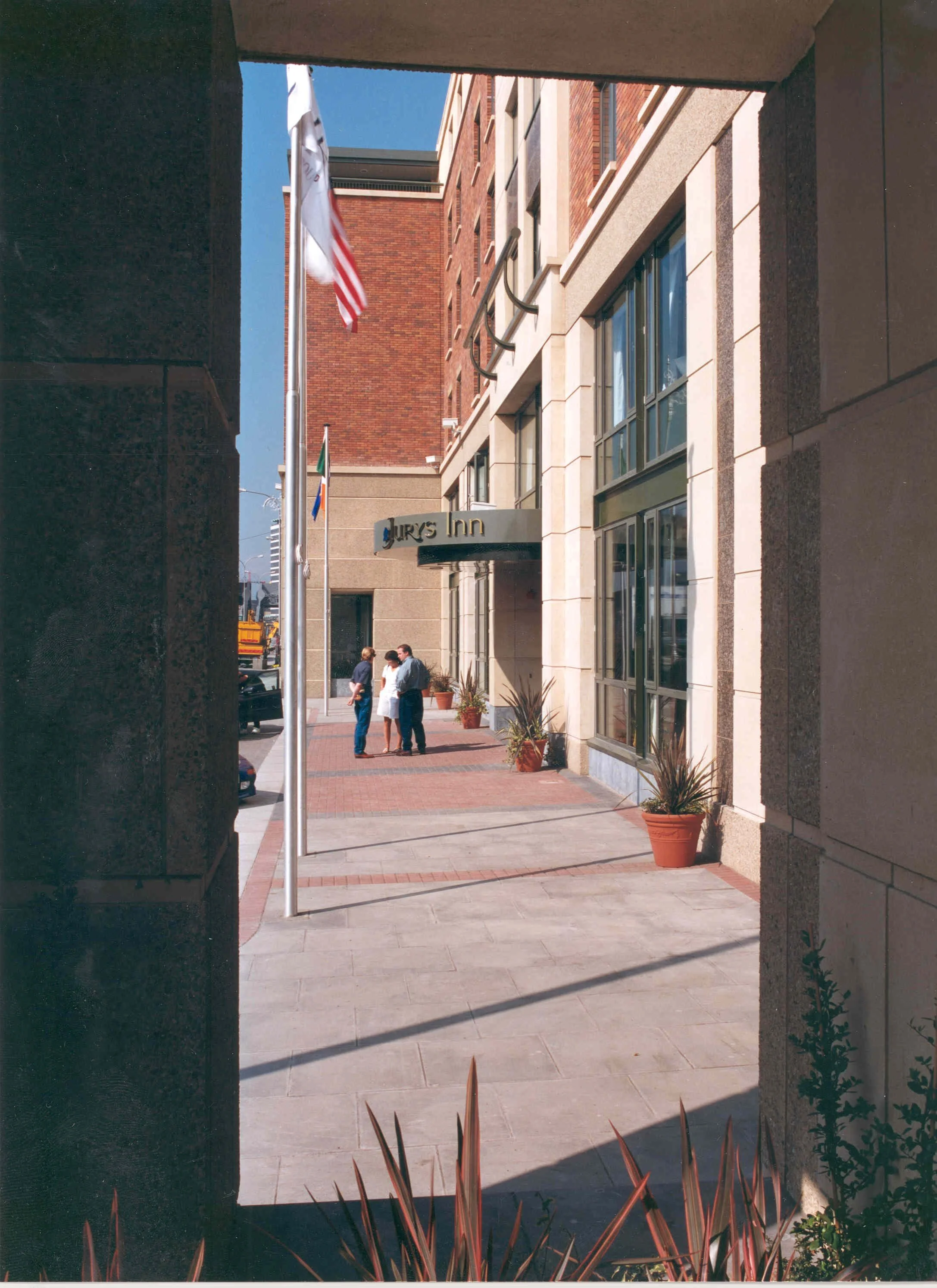 View of the entrance to a Hilton hotel, with a sign that reads 'HILTON.' Three people are standing and talking outside the hotel, with potted plants and American flags nearby.