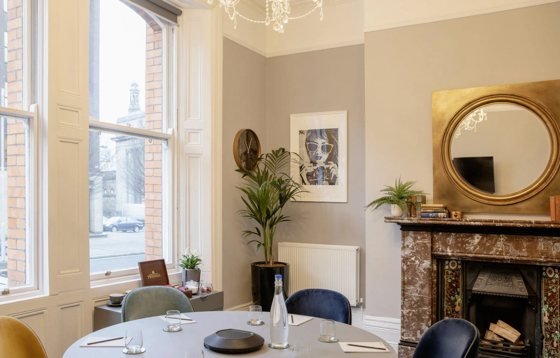 Interior of a dining room with large windows, a chandelier, a fireplace with a mirror above it, a framed artwork, houseplants, and a round dining table set with glasses, a water bottle, and plates.